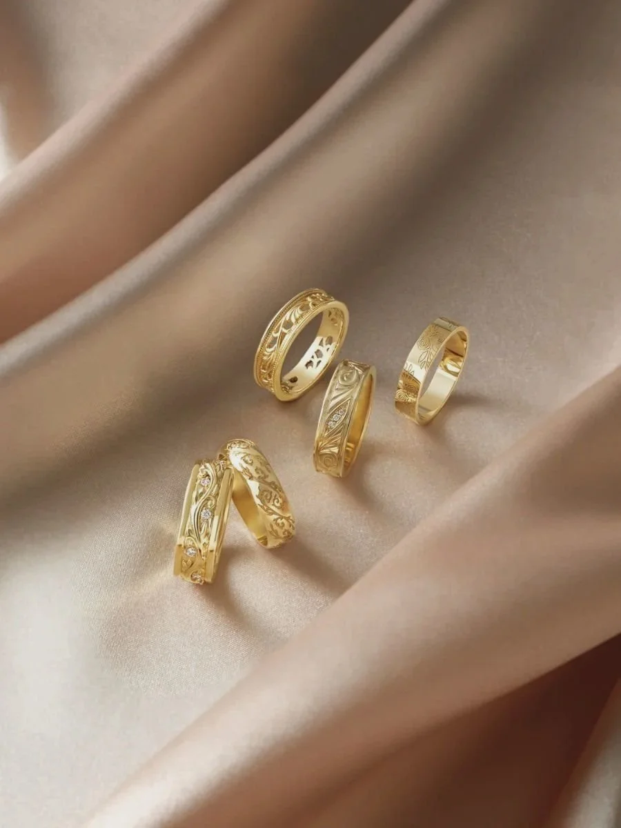 Five gold wedding bands with engraved and decorative details displayed on beige fabric.