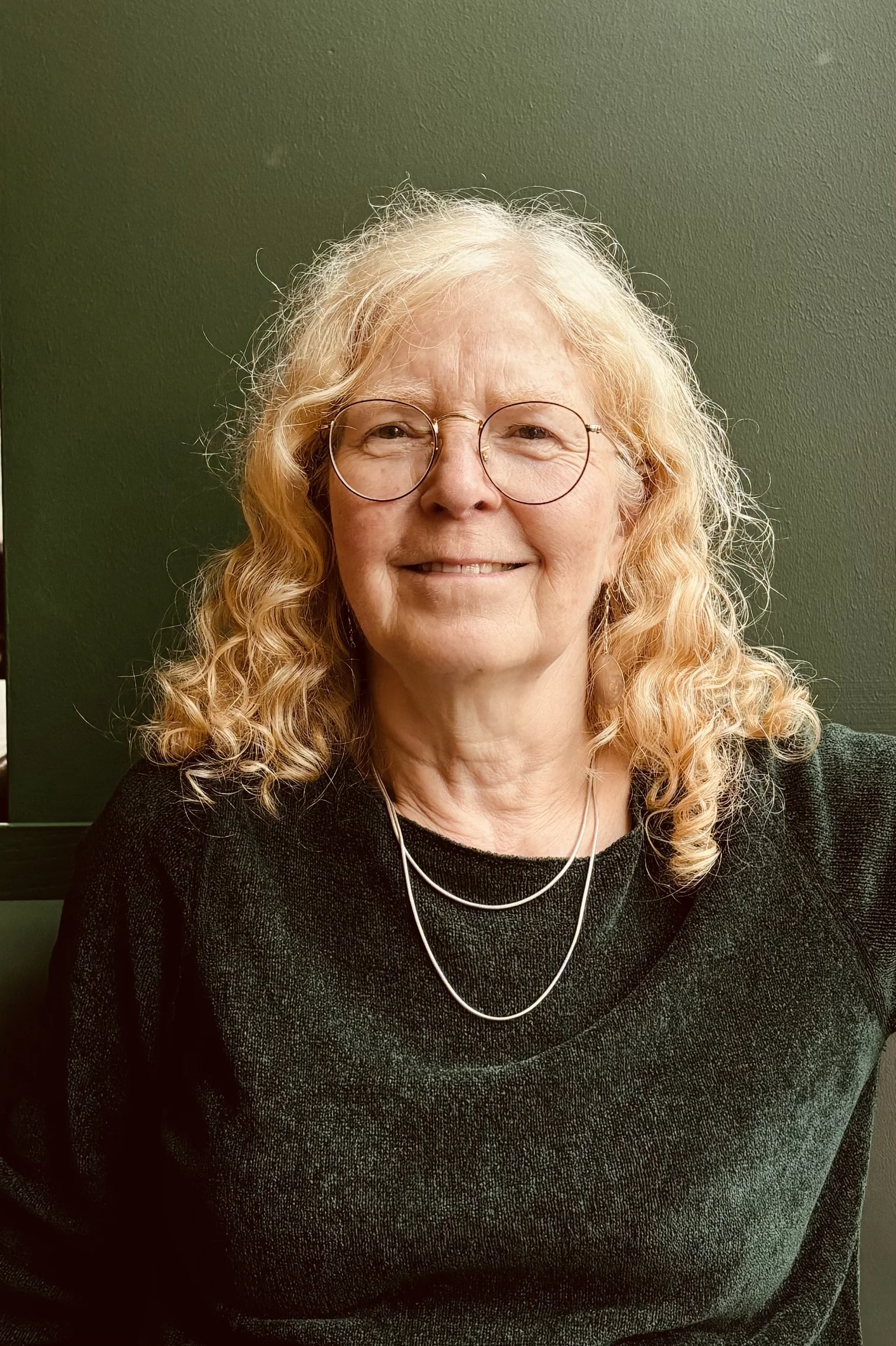 A smiling woman with curly red hair wearing glasses, a green top, and layered necklaces, sitting against a dark green background.