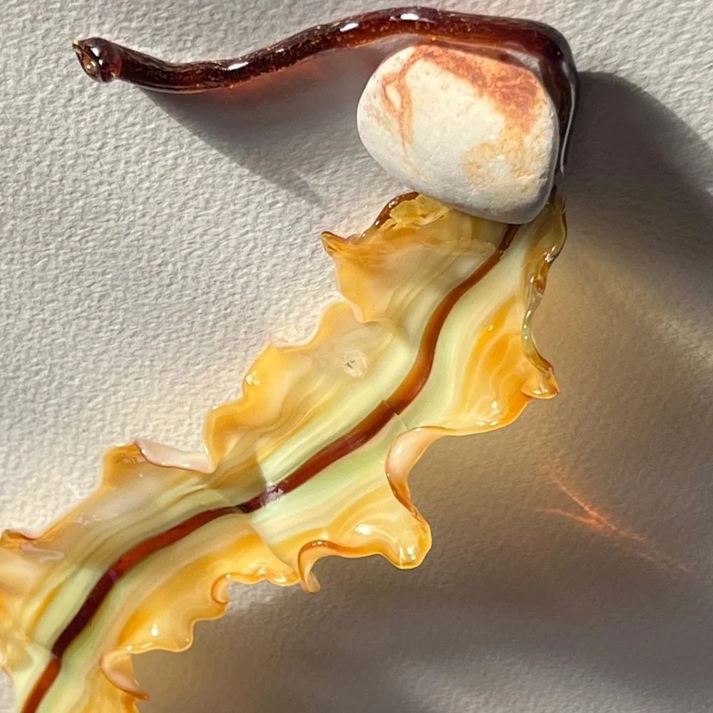 Close-up of an ornate glass decoration resembling a plant stem with wavy edges, along with a small rounded stone or shell at the bottom, on a textured surface.