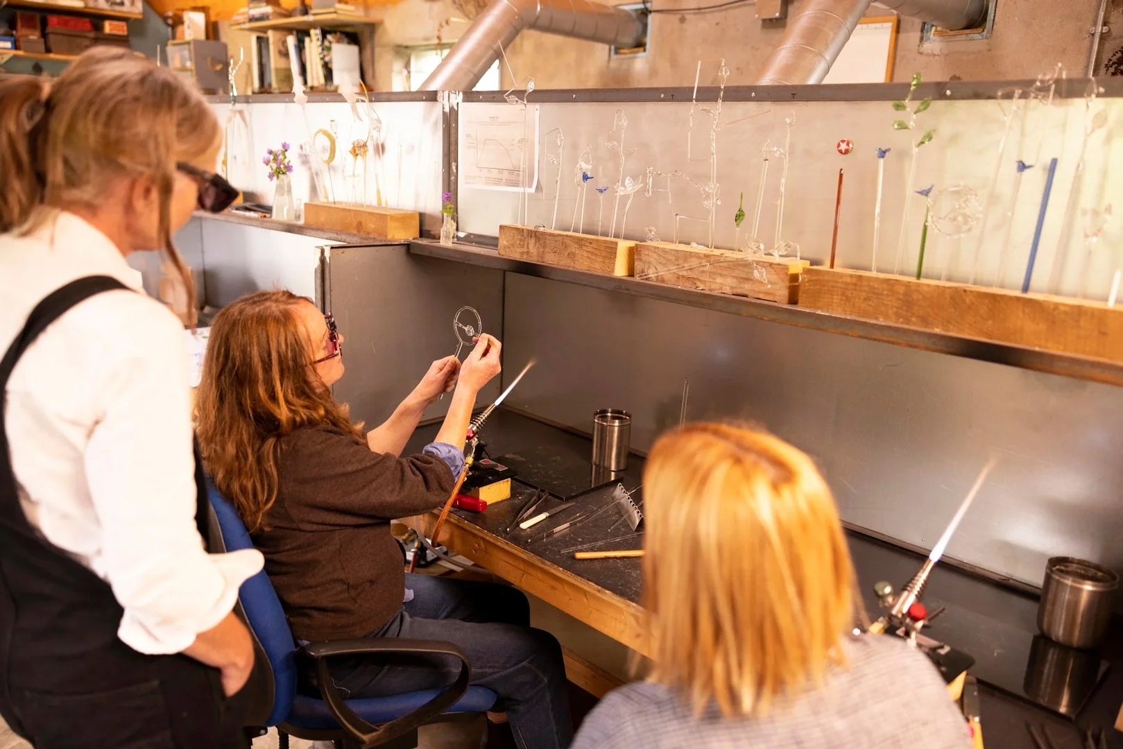Several women working on glassblowing art at a workshop, with glass sculptures on the work surface and shelves.
