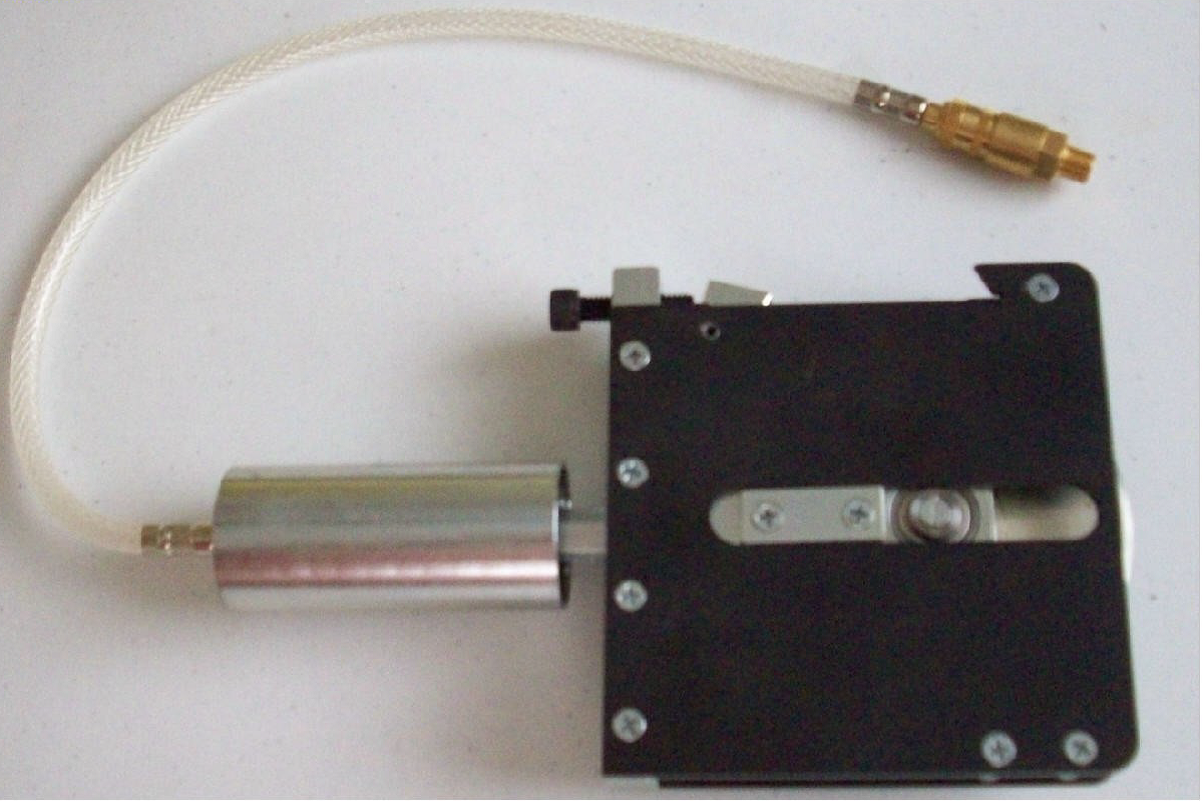 A mechanical device with a black rectangular base, a cylindrical metallic component, and a white braided cable with gold connectors.