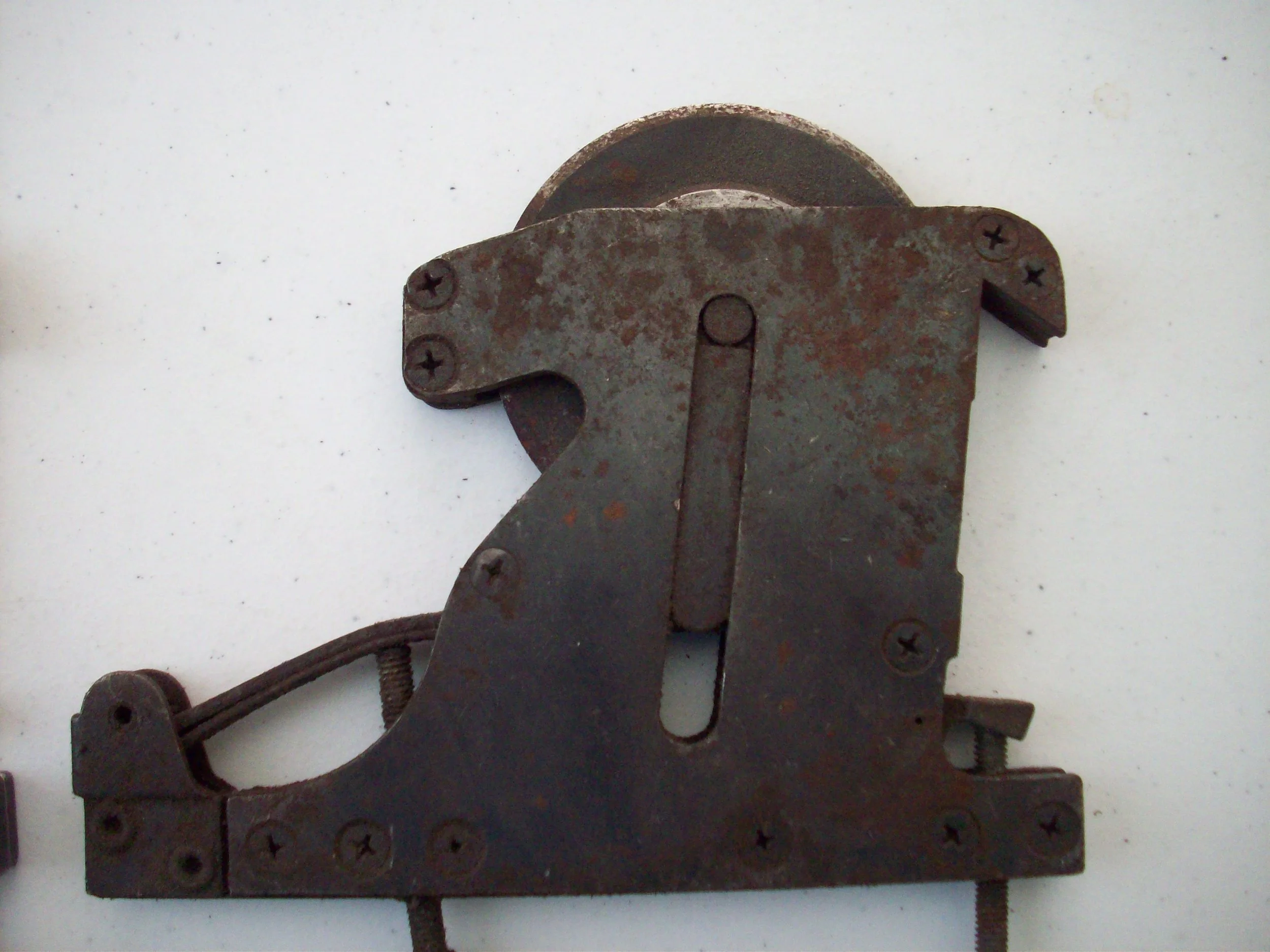 A rusty metal mechanical device with screws, a wheel, and a metal arm, possibly part of a machine or tool, mounted on a white surface.