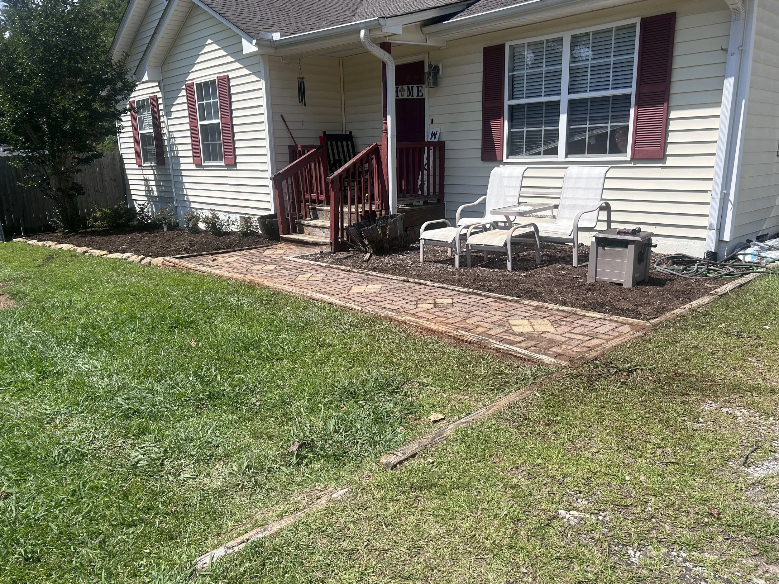Backyard patio area with brick pathway, two white outdoor chairs, and a small gray toolbox against the house with white vinyl siding and maroon shutters. Small garden beds and a wooden fence are visible.