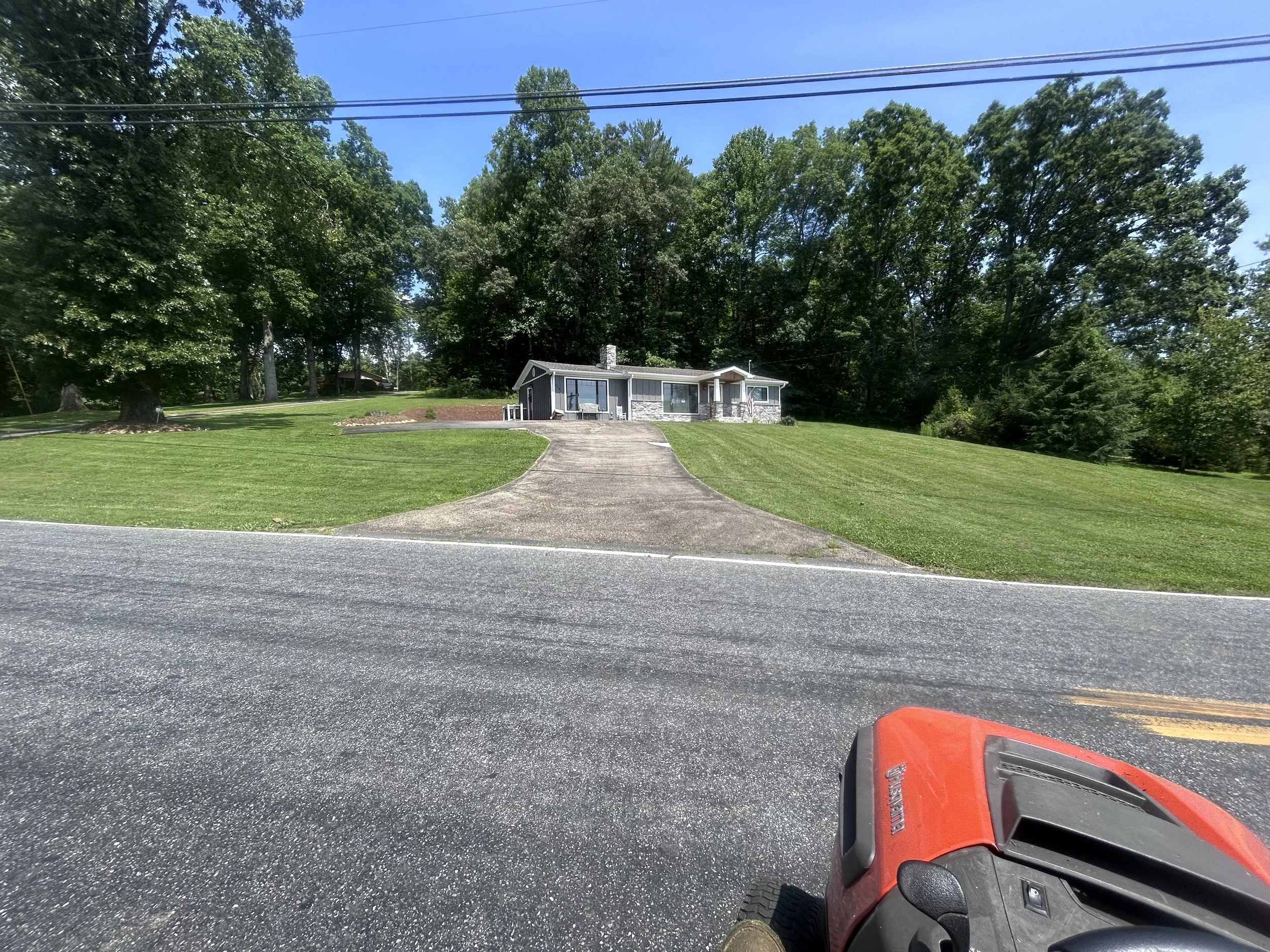 A house on a hill with a paved driveway, lush green grass, and trees in the background, viewed from across the street with a red lawn mower in the foreground.