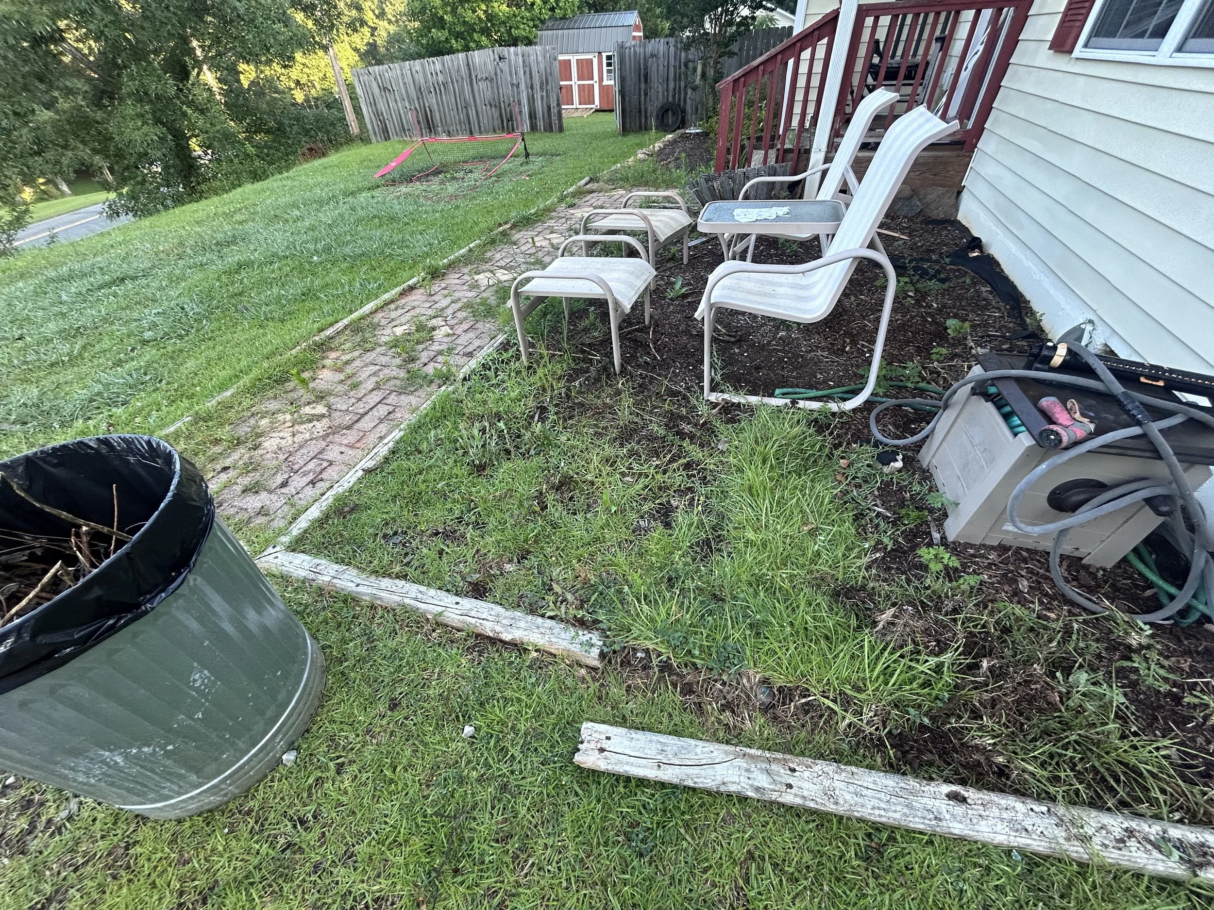 Backyard with a small patio area featuring four white chairs, a small metal table, a black trash can filled with sticks, and a white storage box. The yard has grass, a brick pathway, and a grassy hill in the background. There is a red fence, a shed, and some gardening tools near the house.