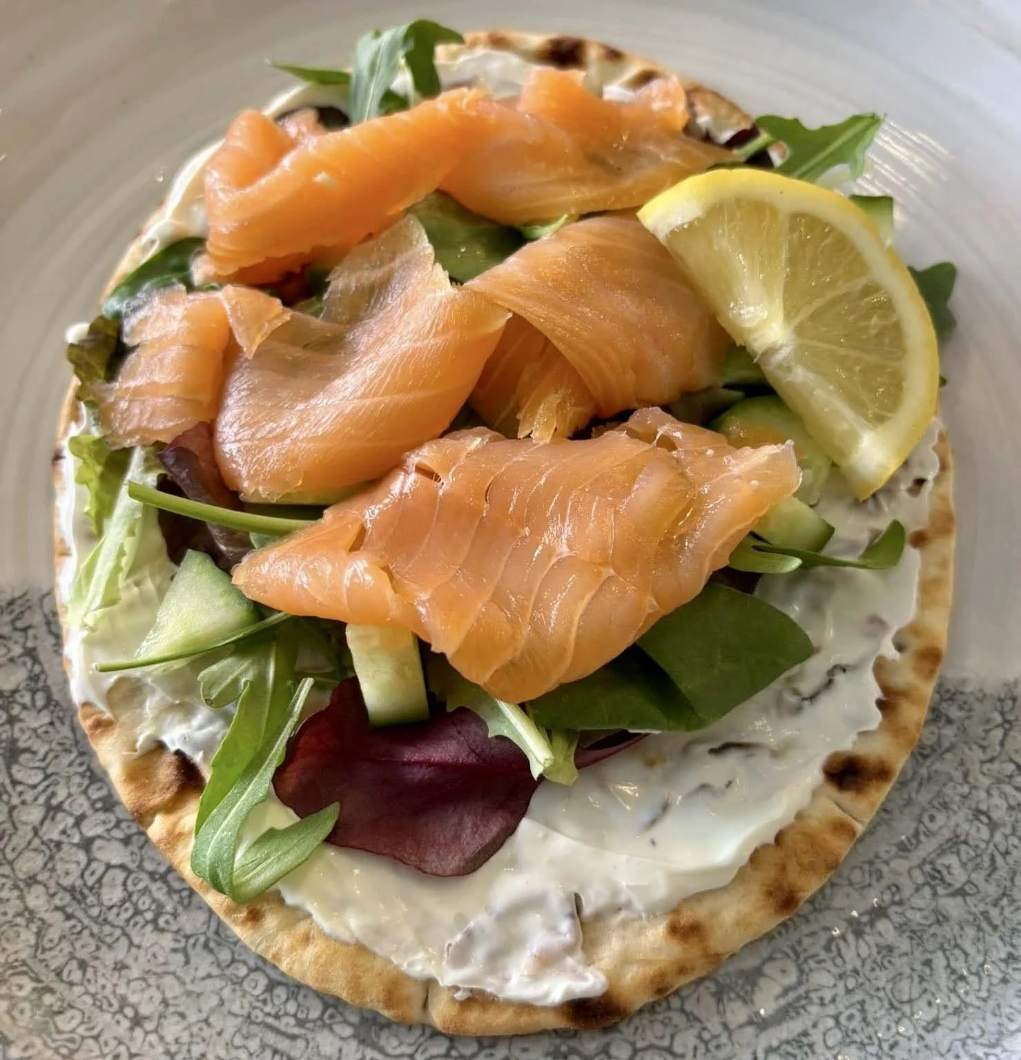 A flatbread topped with smoked salmon, mixed greens, cucumber, and a lemon wedge, served on a ceramic plate.