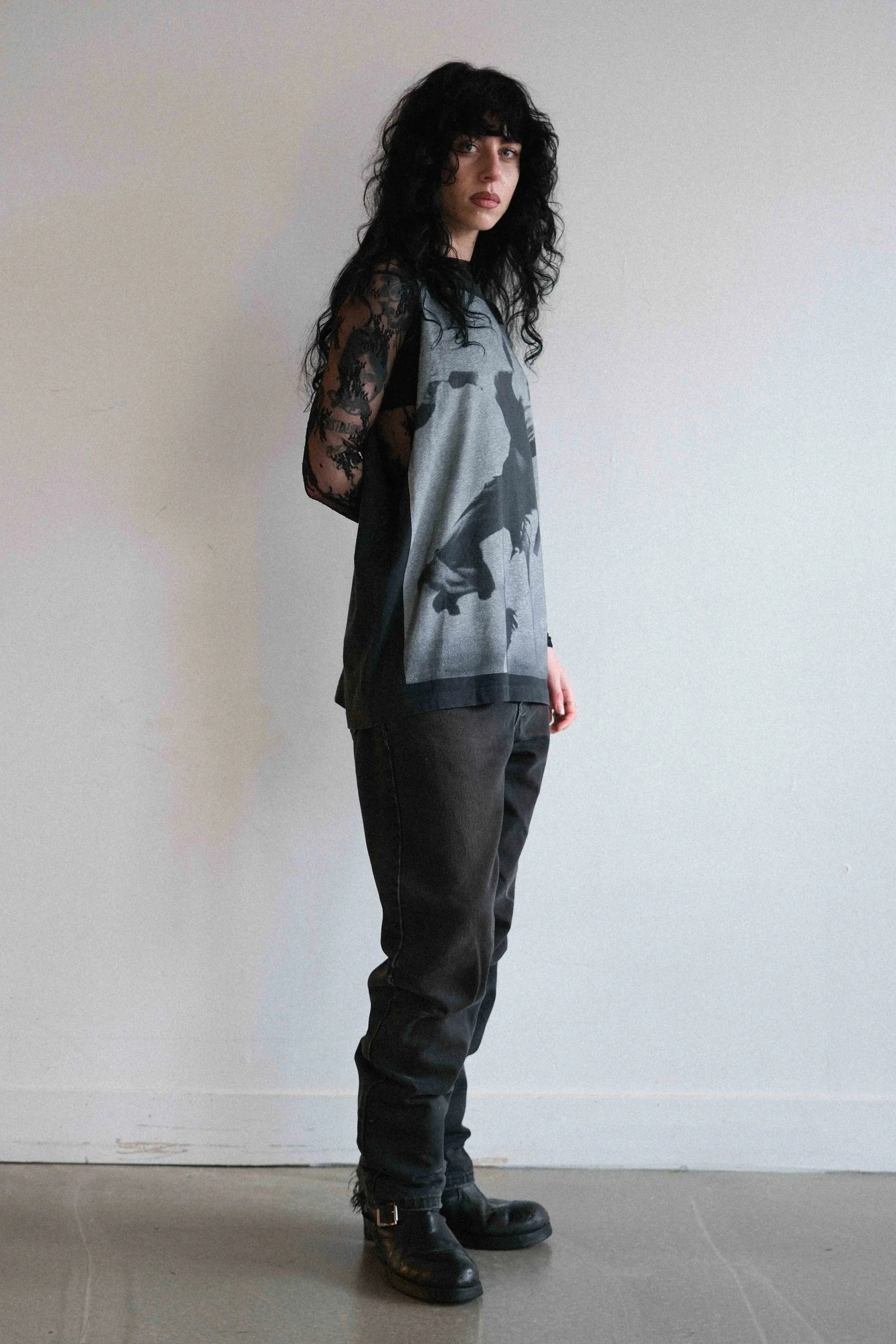 A woman with black curly hair wearing a layered black lace long-sleeve top under a gray graphic T-shirt, black pants, and black boots standing against a plain white wall.
