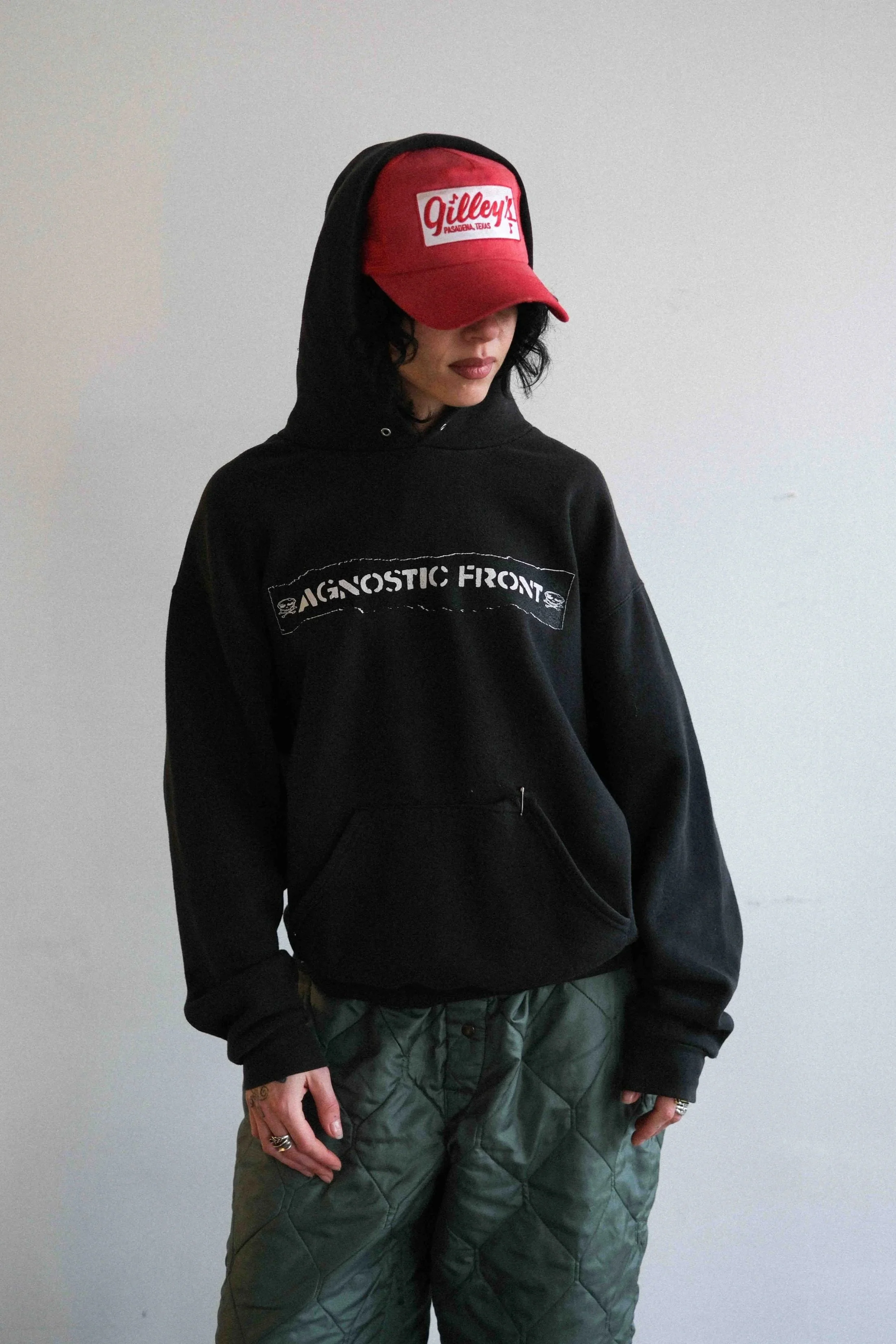 Person wearing a red and white Gilley's Pasadena, Texas cap, black hoodie with 'Ignostic Front' logo, and green quilted pants, standing against a plain wall.