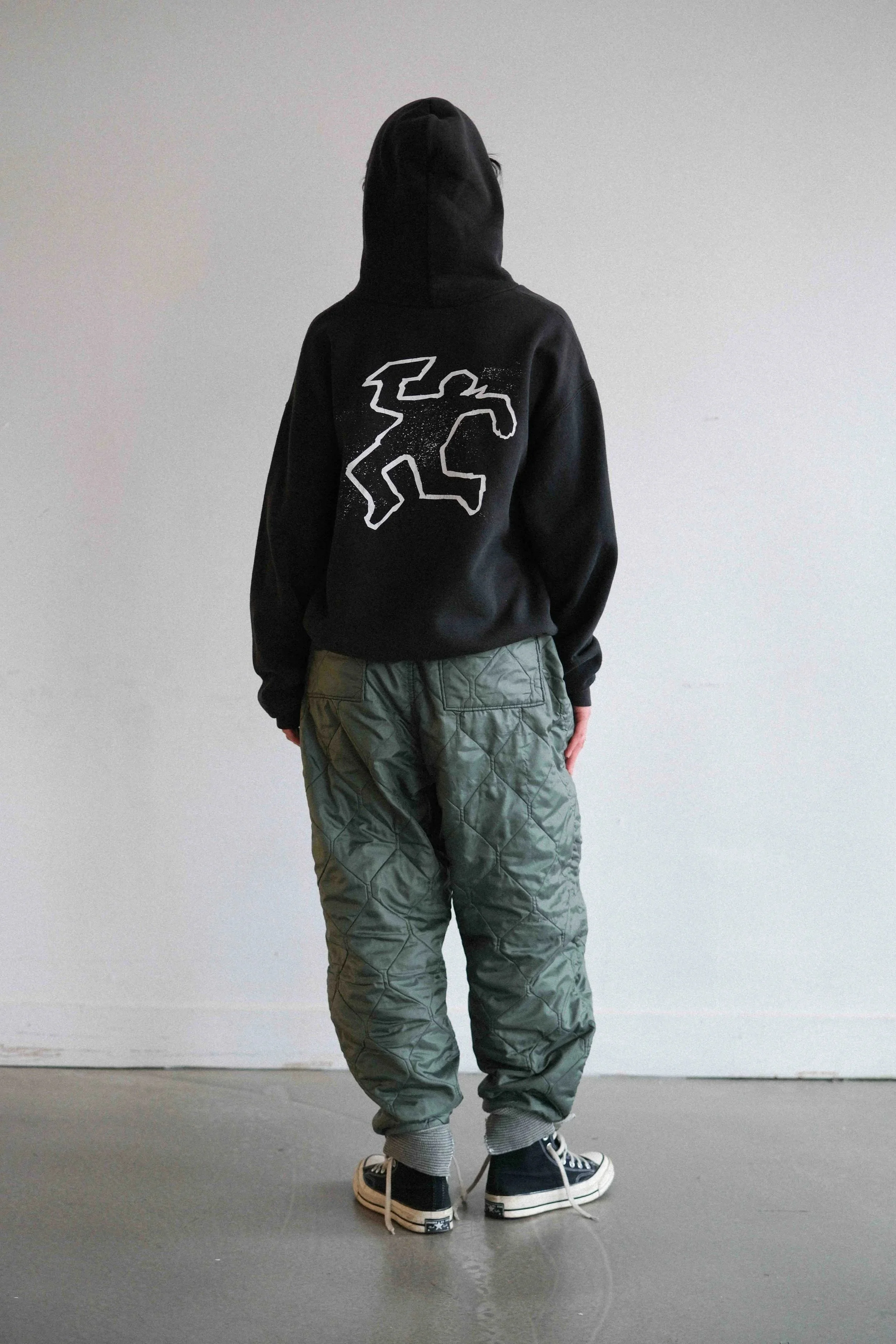 Person wearing a black hoodie with a white graphic on the back, green baggy pants, and black sneakers standing against a white wall.