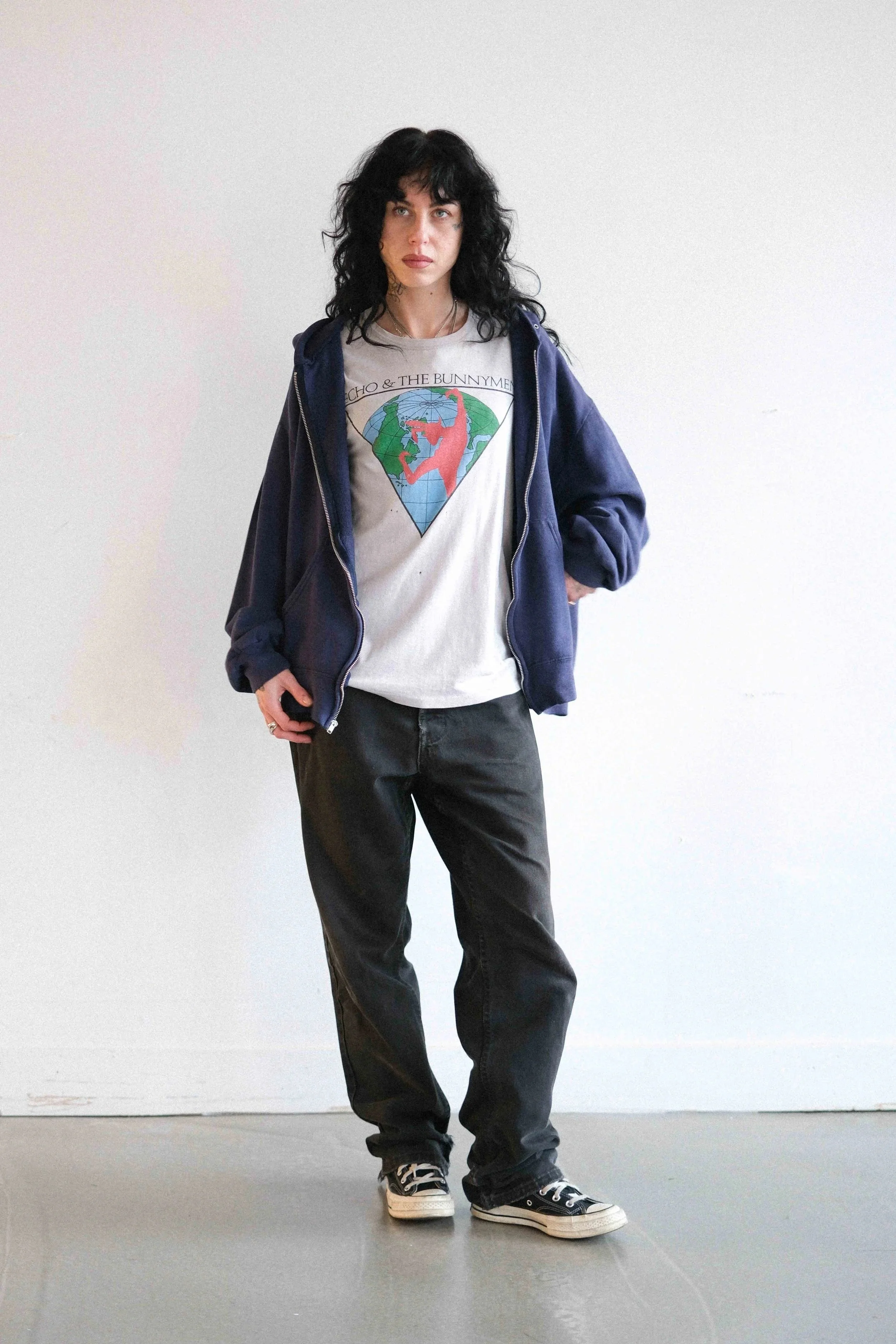 Young woman with black hair and casual attire standing against a plain white wall, wearing a graphic t-shirt, navy hoodie, black jeans, and sneakers.