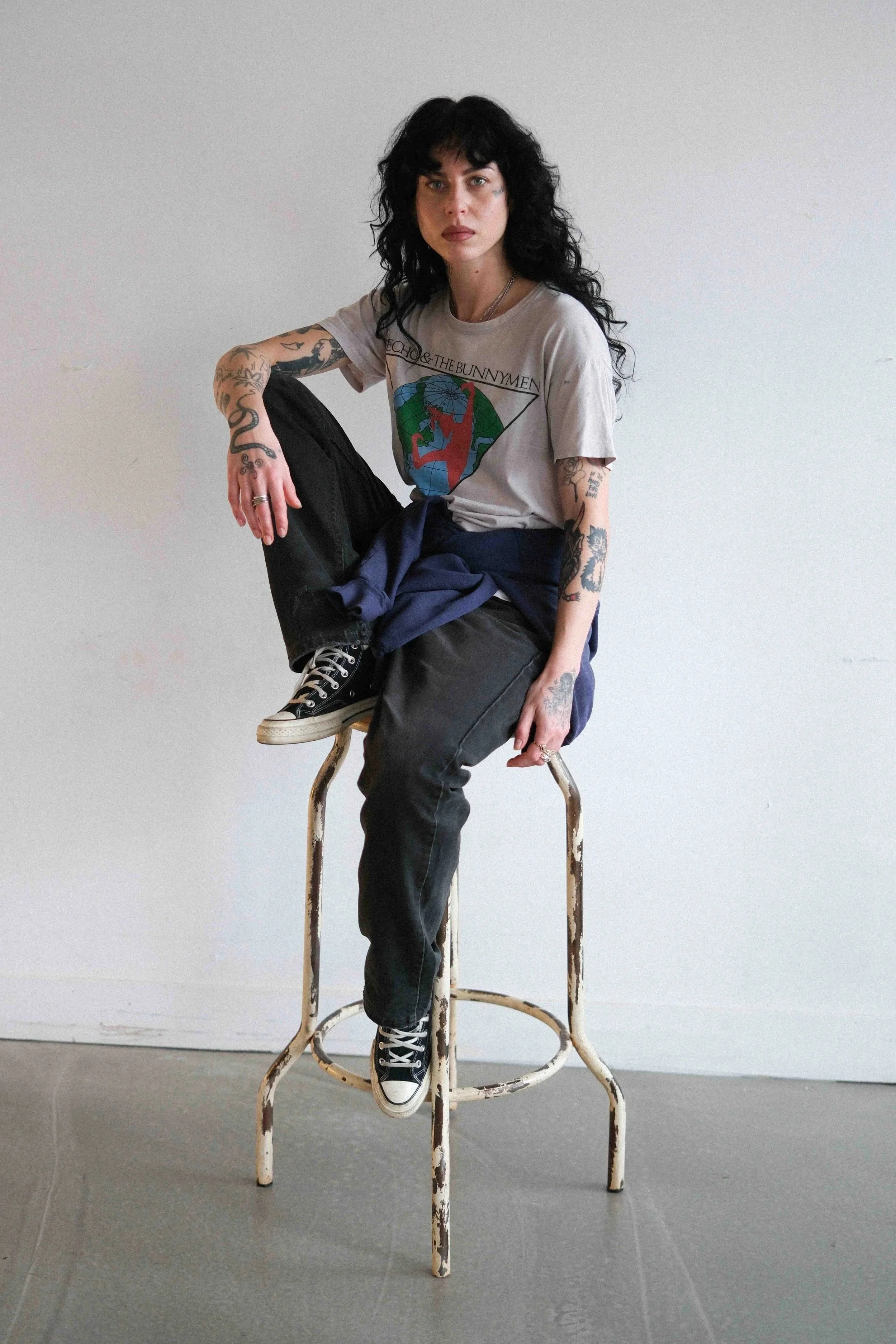 A woman with black curly hair and tattoos, dressed in a vintage T-shirt and jeans, sitting on an old worn barber's stool with one foot on the seat and the other on the rung, against a plain white wall.