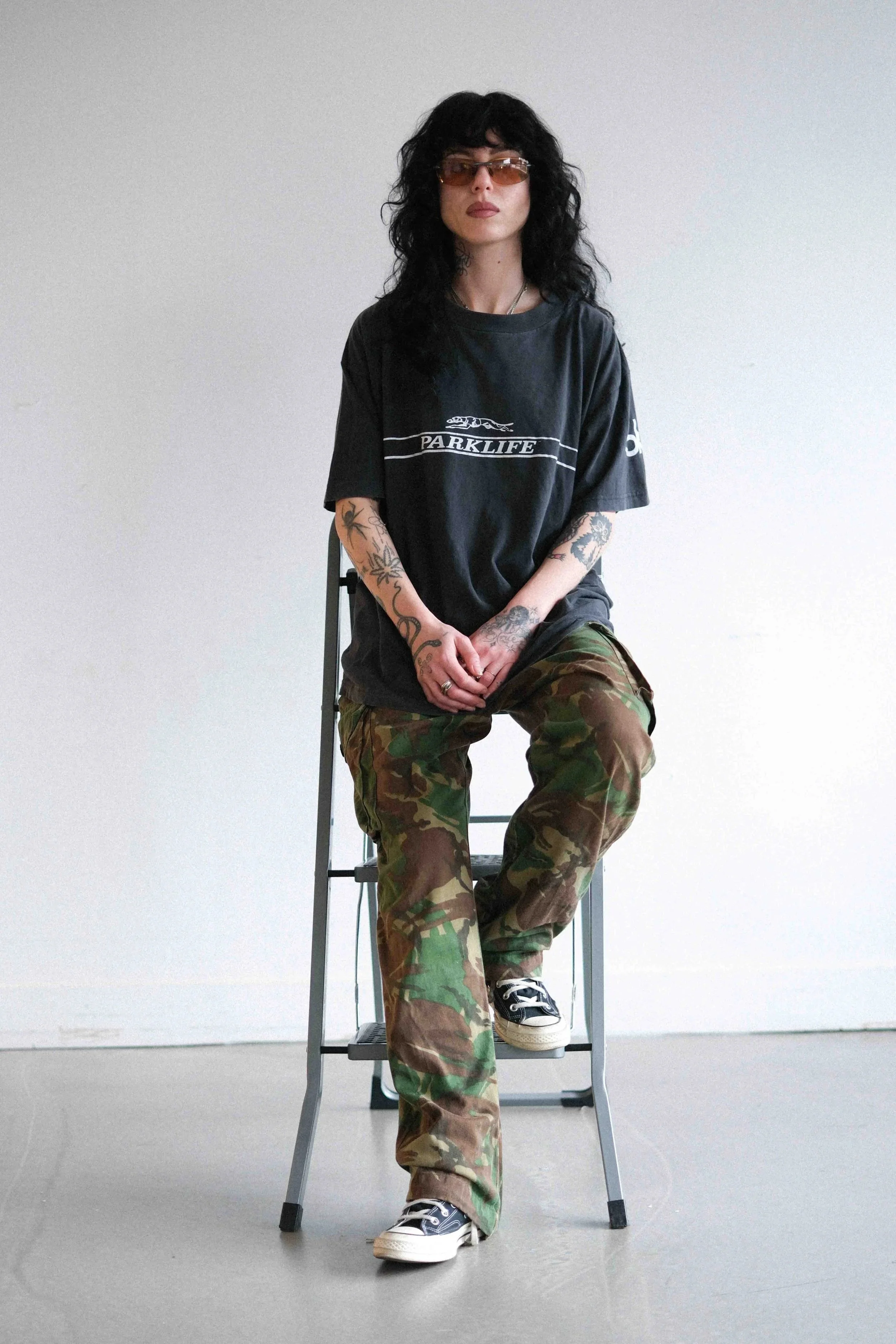 A woman with black curly hair, wearing sunglasses, a black T-shirt with 'PARKLIFE' and a graphic of a car, camouflage pants, and black sneakers, sitting on a stool against a plain white background.