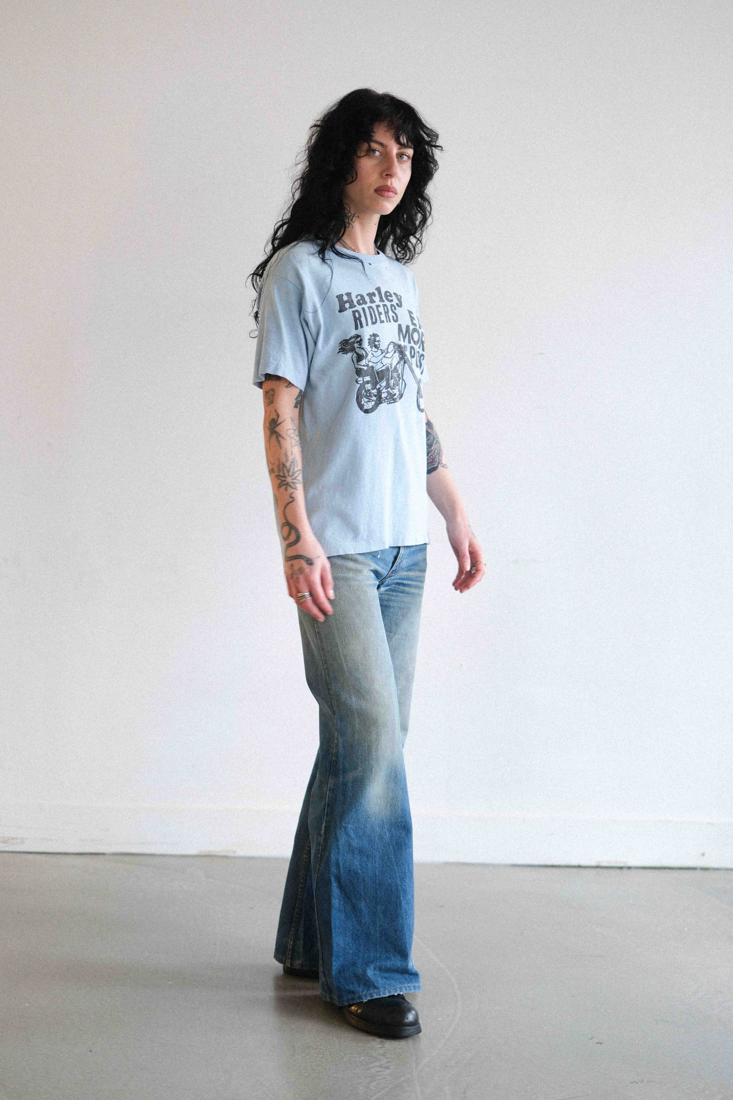 A woman with curly black hair and tattoos on her arms, standing against a plain white wall, wearing a light blue vintage Harley-Davidson T-shirt and flared jeans.
