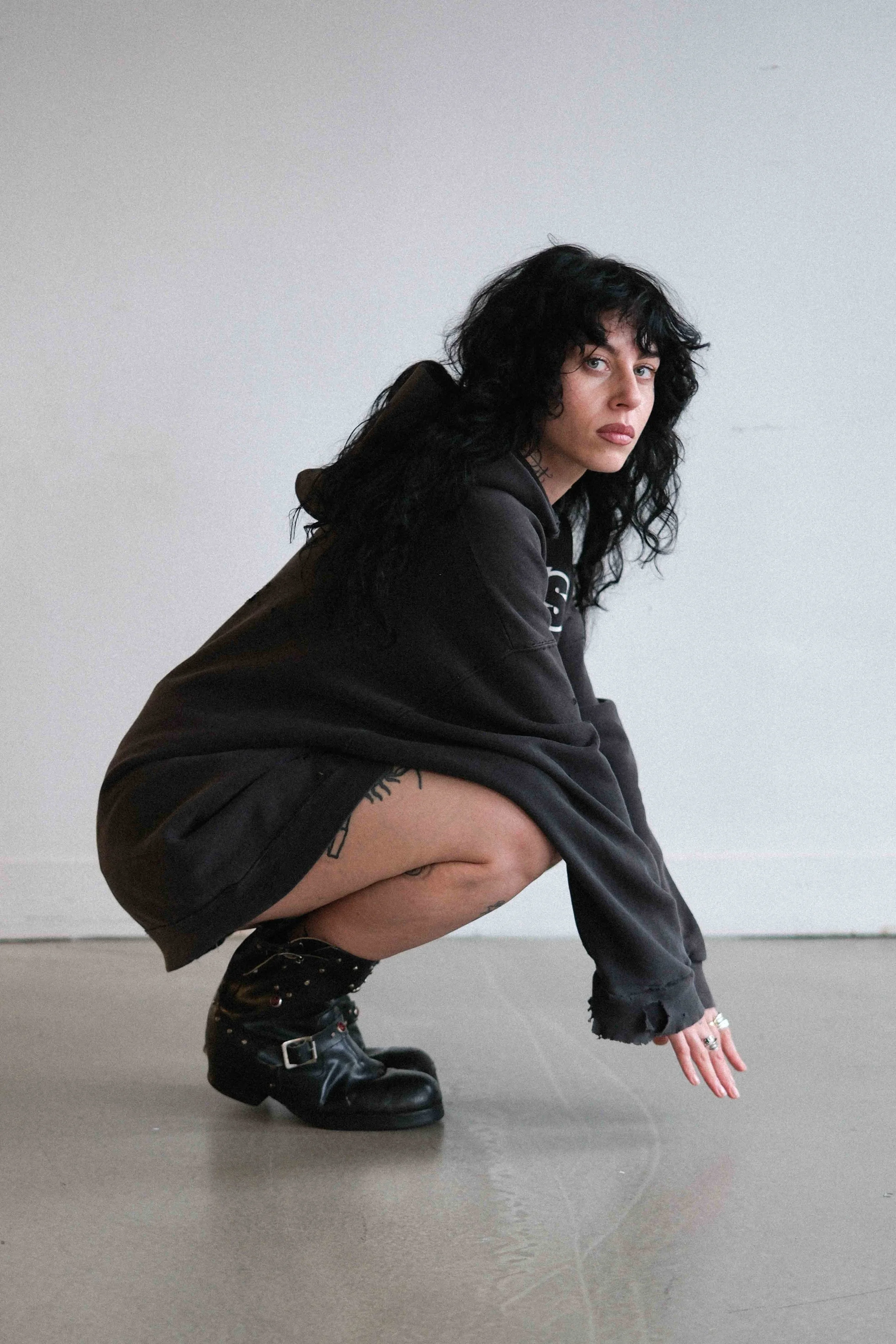 A woman with black curly hair squatting on the floor, wearing a black hoodie, shorts, and black combat boots, against a plain white background.