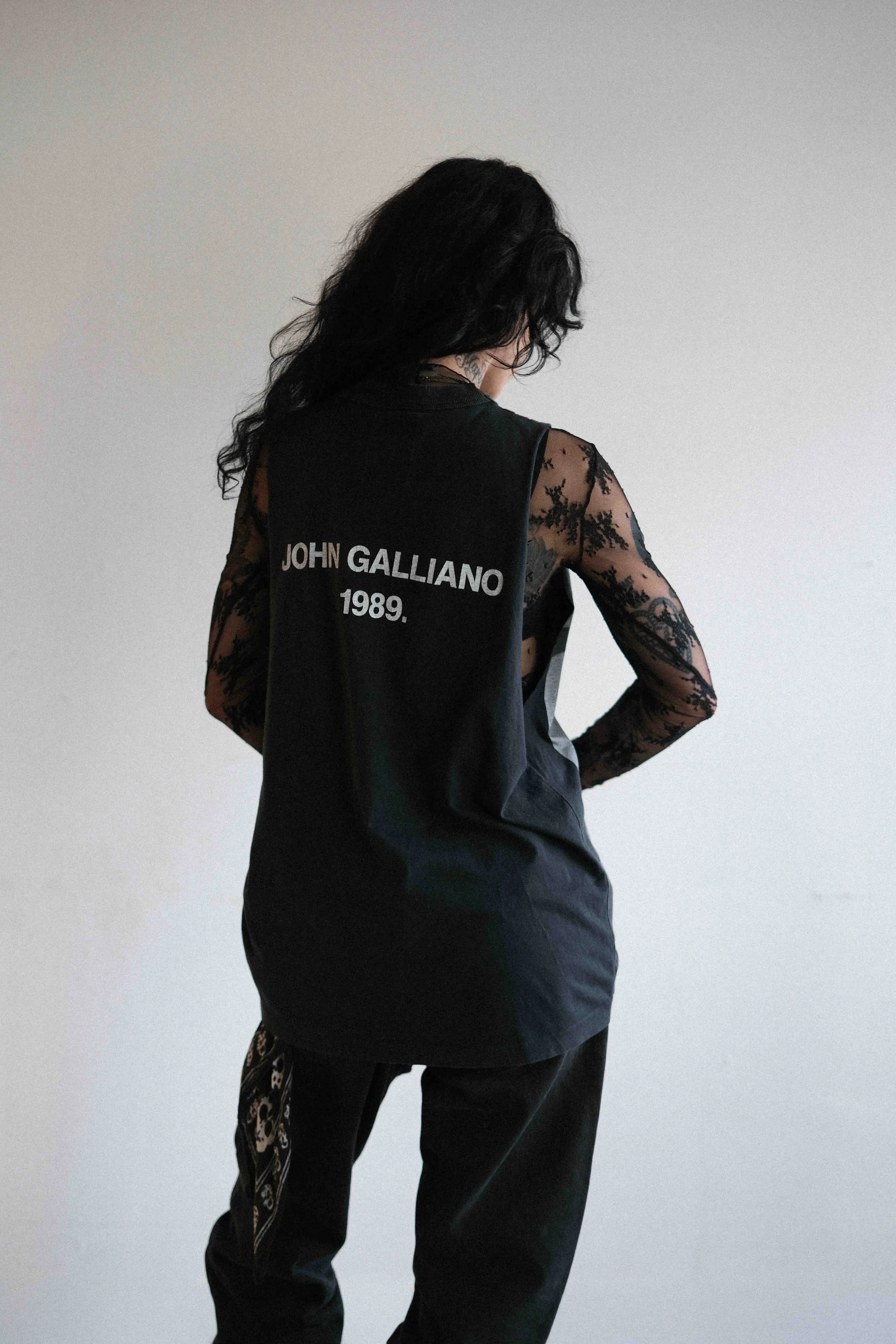 A person with long dark hair, wearing a black sleeveless top with lace long sleeves underneath, with text reading 'JOHN GALLIANO 1989' on the back, standing against a plain light-colored wall.