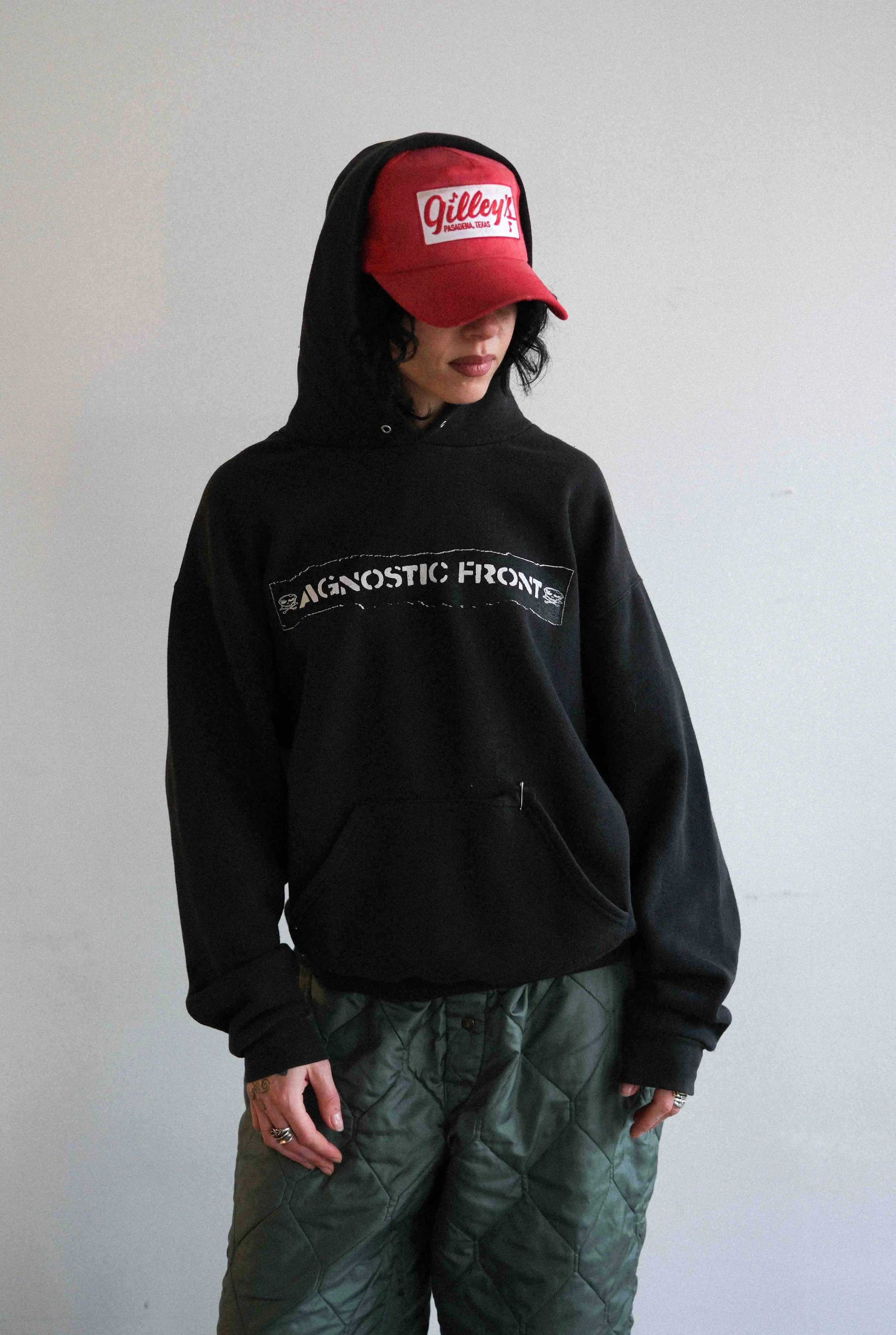 A woman with dark hair wearing a black hoodie, a red cap with white text, and green quilted pants, standing against a plain light gray background.