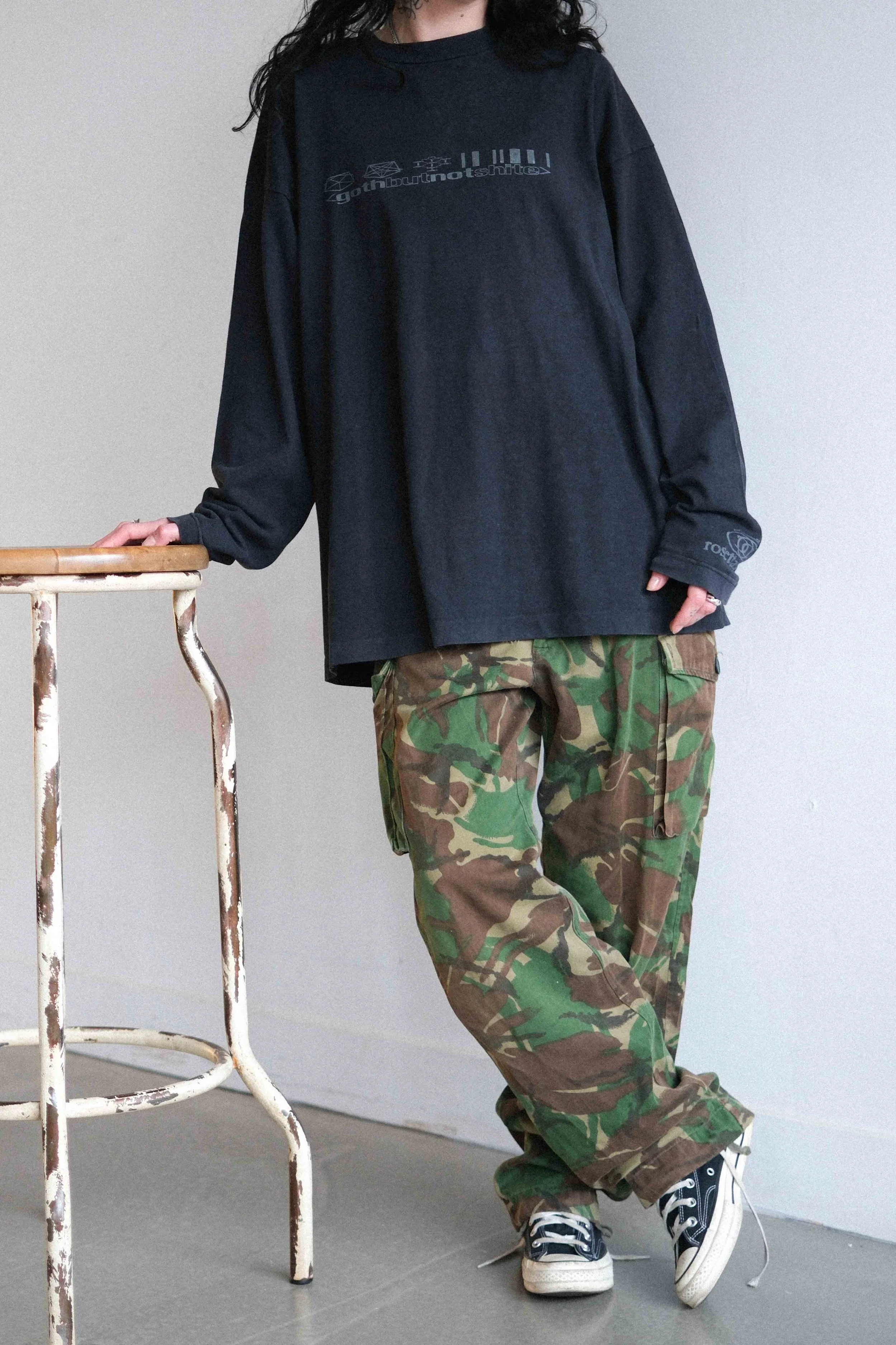 Person wearing a oversized black shirt and camouflage cargo pants, standing next to a rustic, distressed metal stool.