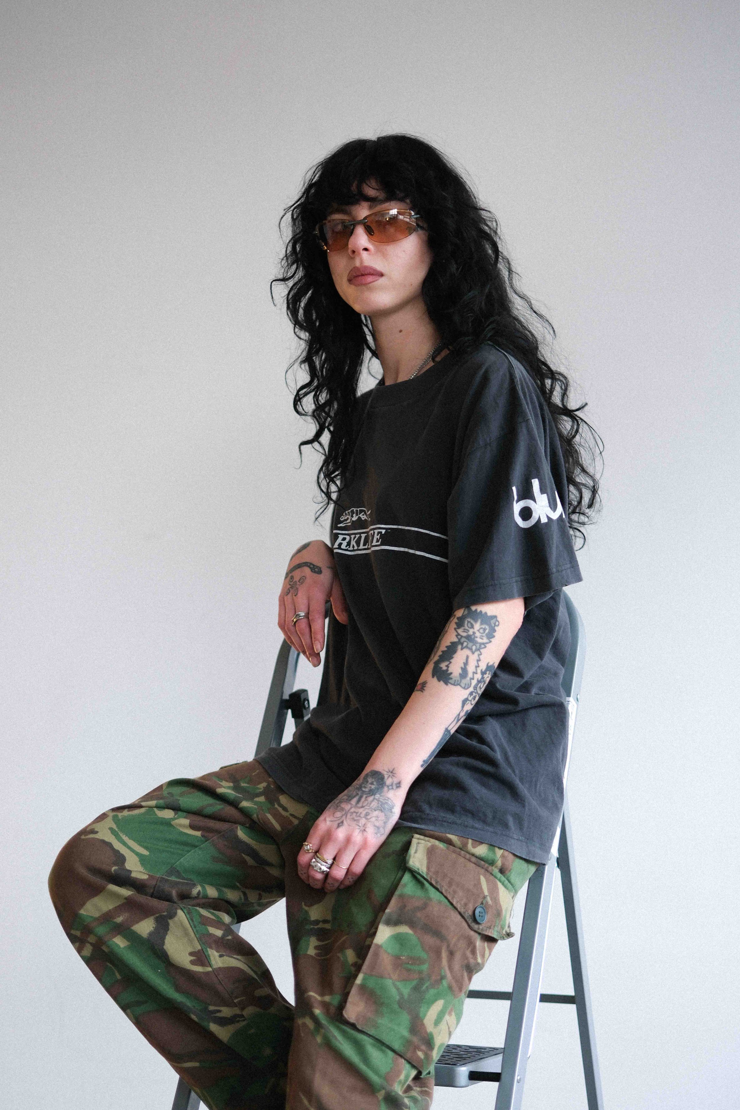 A woman with long black curly hair and tattoos, wearing sunglasses, a black T-shirt, and camouflage pants, sitting on a step stool against a plain white background.