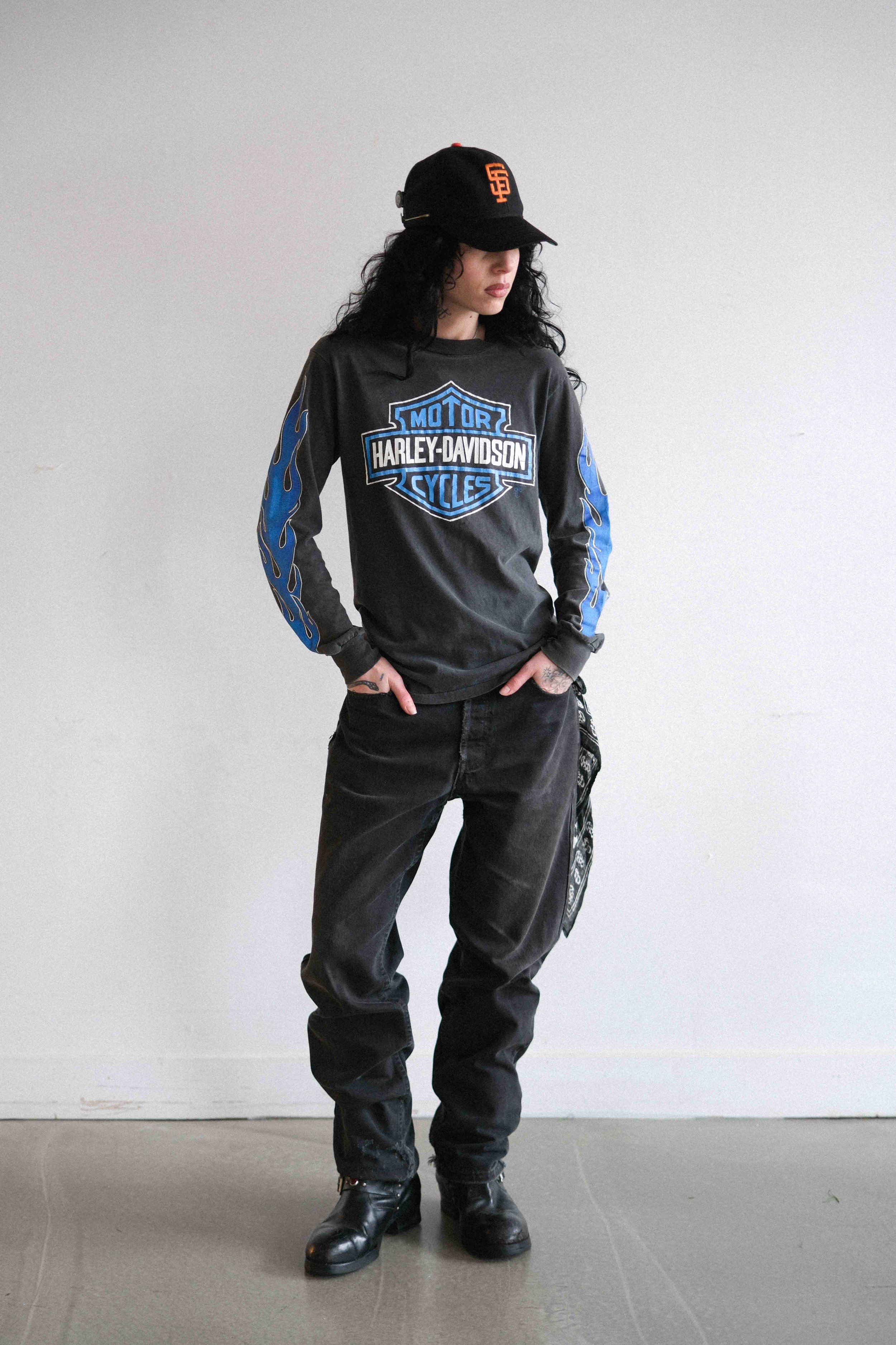 A woman with curly black hair wearing a black Harley-Davidson sweatshirt with blue flames on the sleeves, black pants, black boots, and a black cap with orange logo, standing against a plain white background.