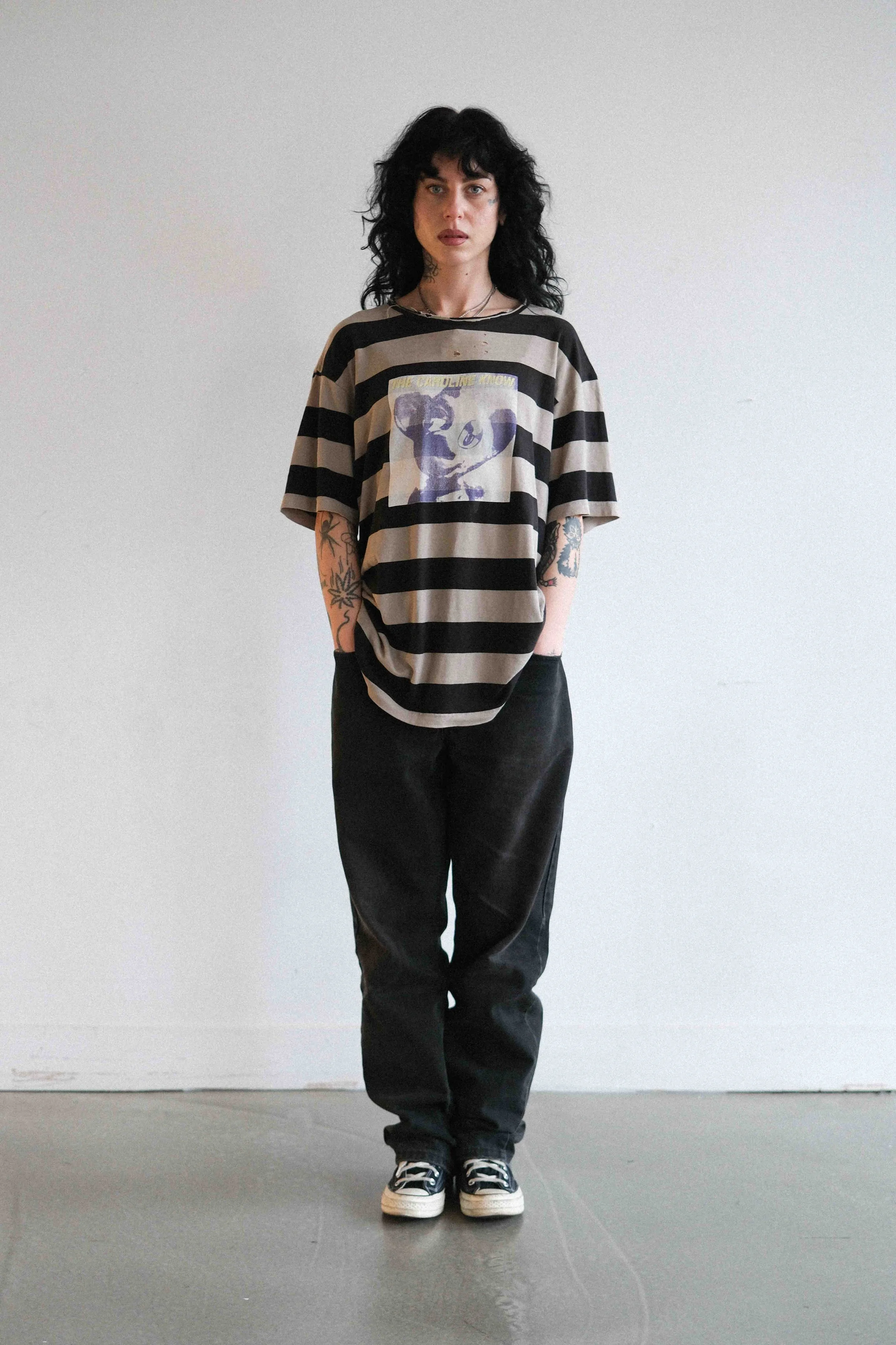 A woman with black, curly hair wearing a striped T-shirt with a graphic of a raccoon and the text "The Caroline Know," black pants, and black and white sneakers, standing against a plain white wall.