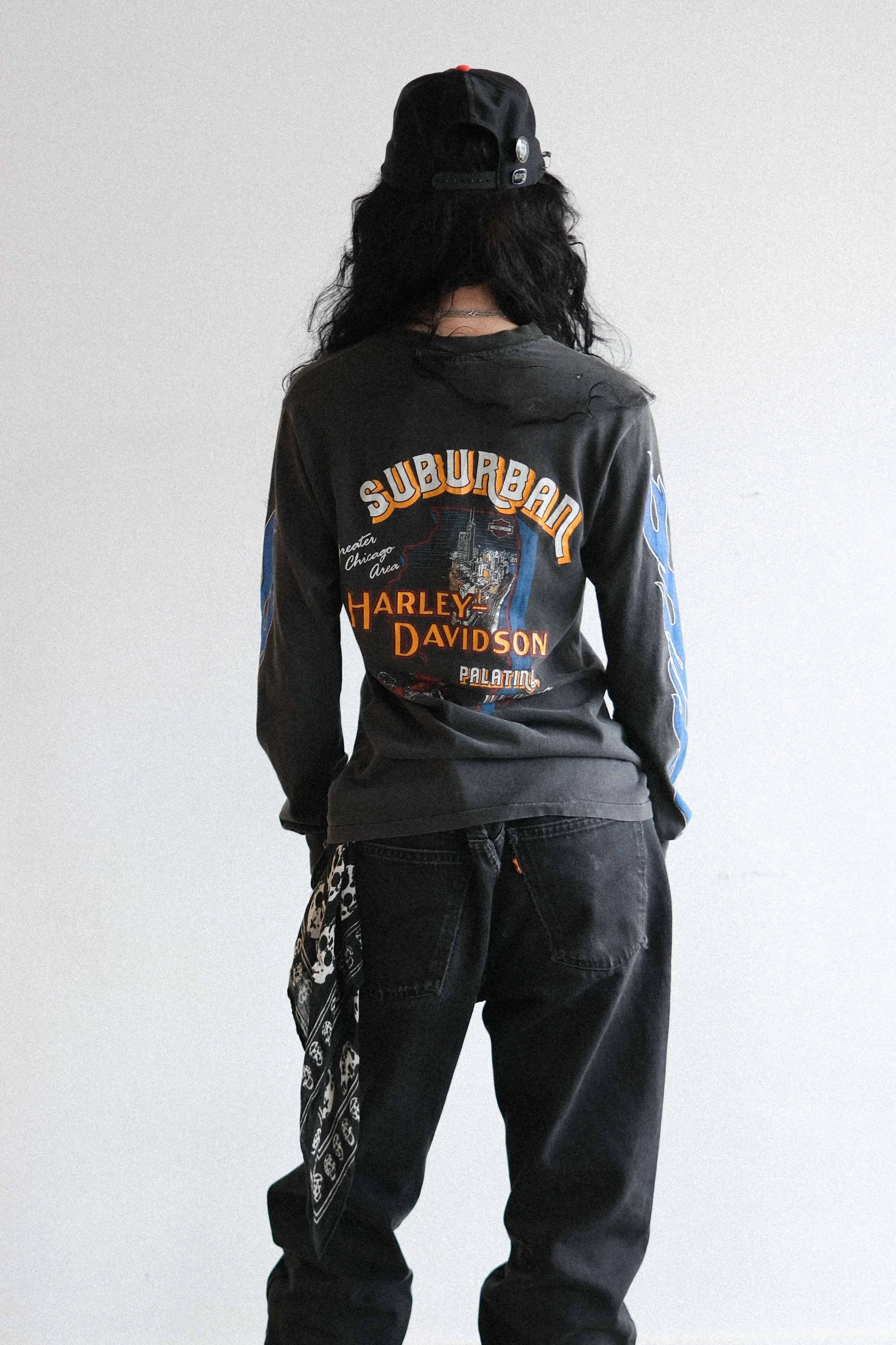 Back view of a person wearing a black Harley Davidson sweatshirt, black jeans, a black baseball cap worn backward, and a black and white bandana hanging from their pocket, standing against a plain wall.