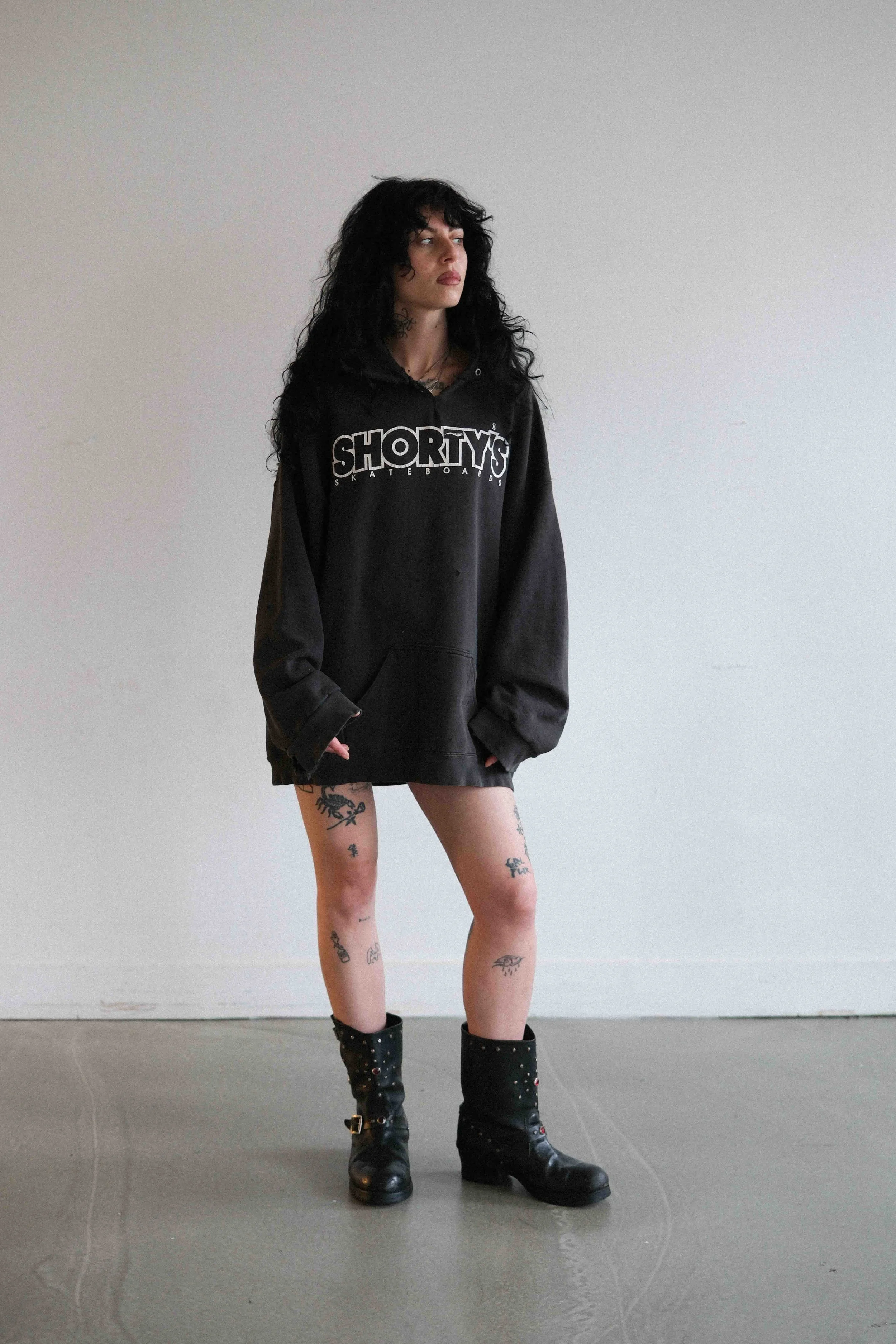 A woman with long black curly hair stands against a plain white wall. She is wearing an oversized black hoodie with the word 'SHORTY'S' on it and black boots with studs. She has visible tattoos on her legs and a confident pose.