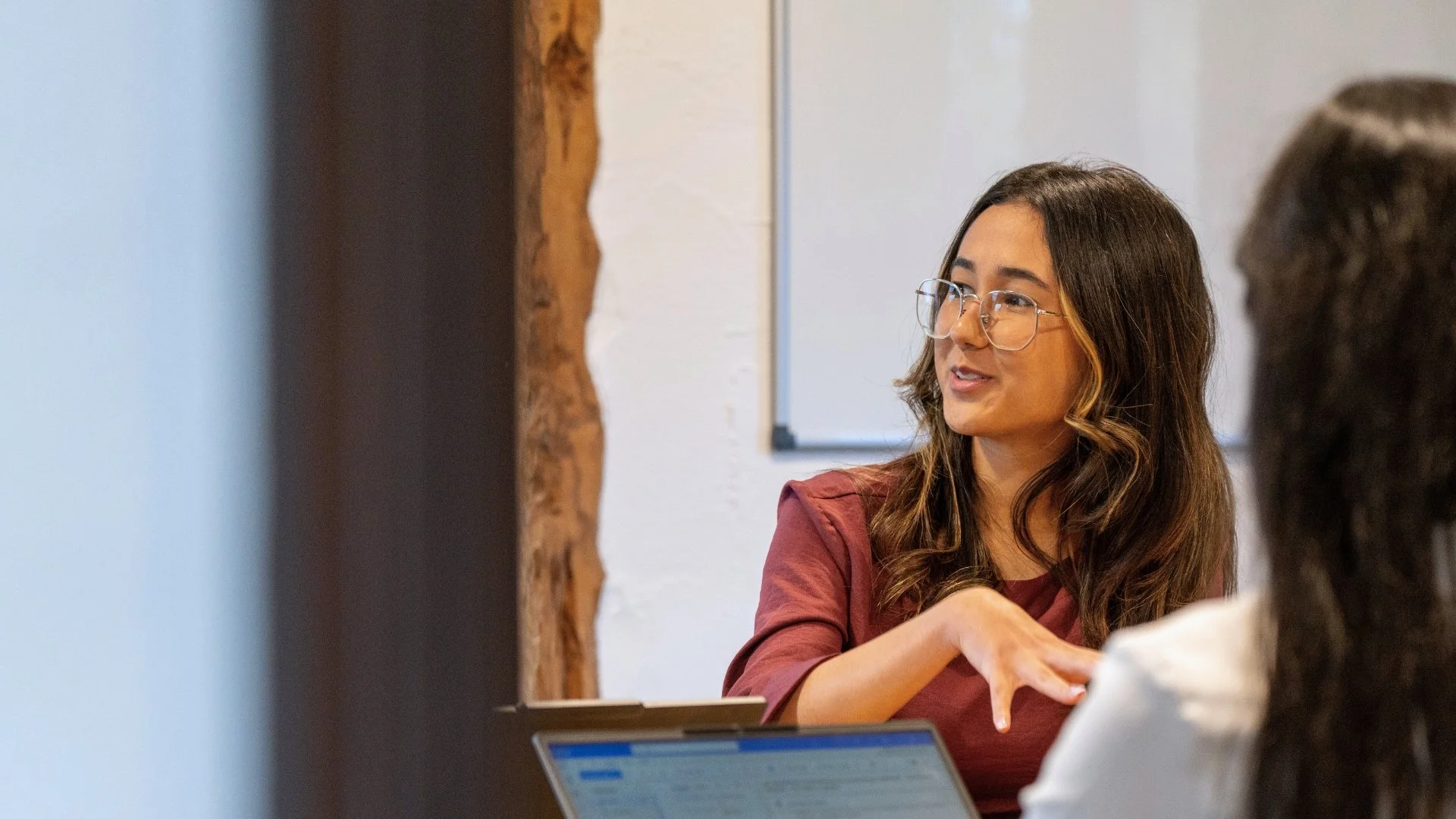 A woman with long brown hair, glasses, and wearing a red shirt is speaking and gesturing during a conversation or meeting. She is engaged and looks focused.