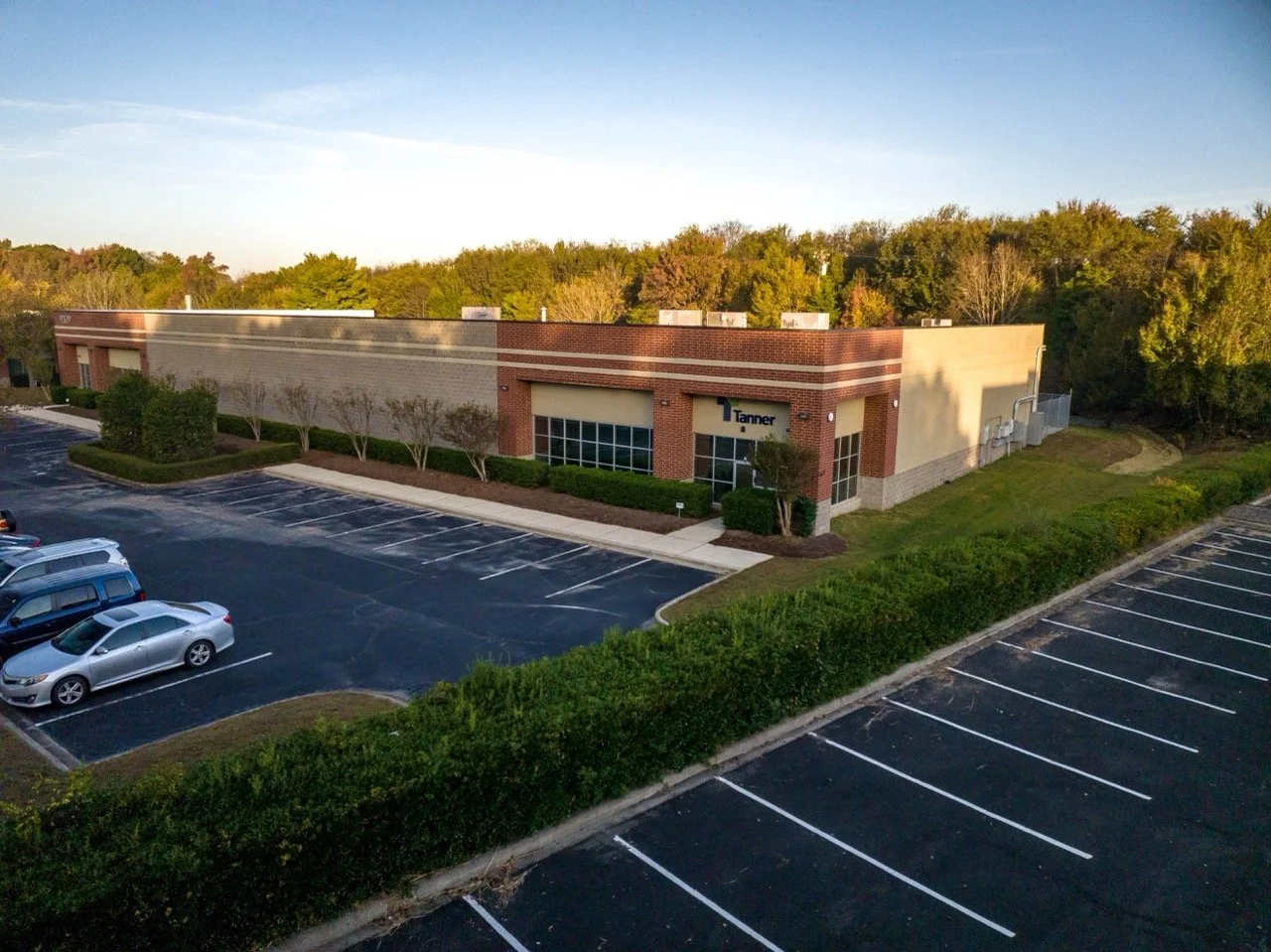 An aerial view of a single-story commercial building with a parking lot, surrounded by trees and greenery, with the logo 'Tanner' on the front.