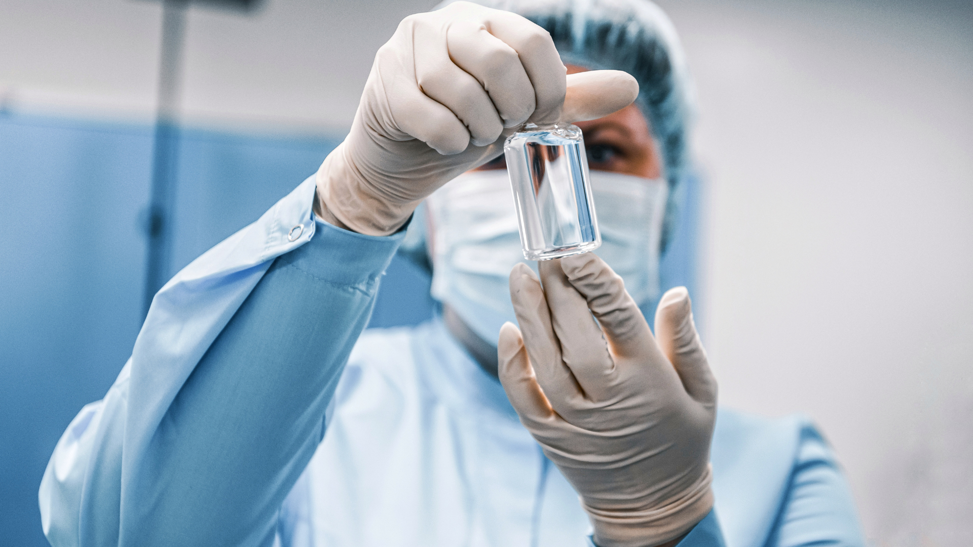 A scientist wearing gloves, a mask, and a hair cap holds a small bottle of clear liquid, examining it closely in a laboratory setting.