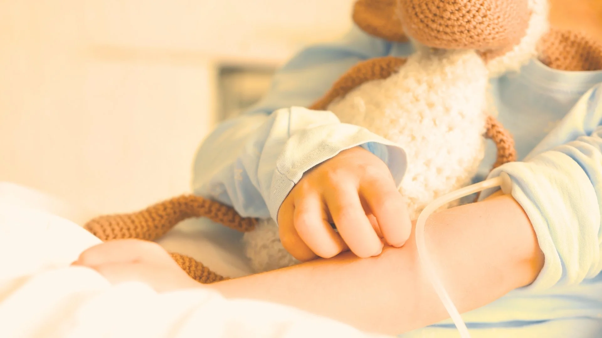 Close-up of a child's hand gripping an adult's finger, with medical IV line attached, in a hospital setting.