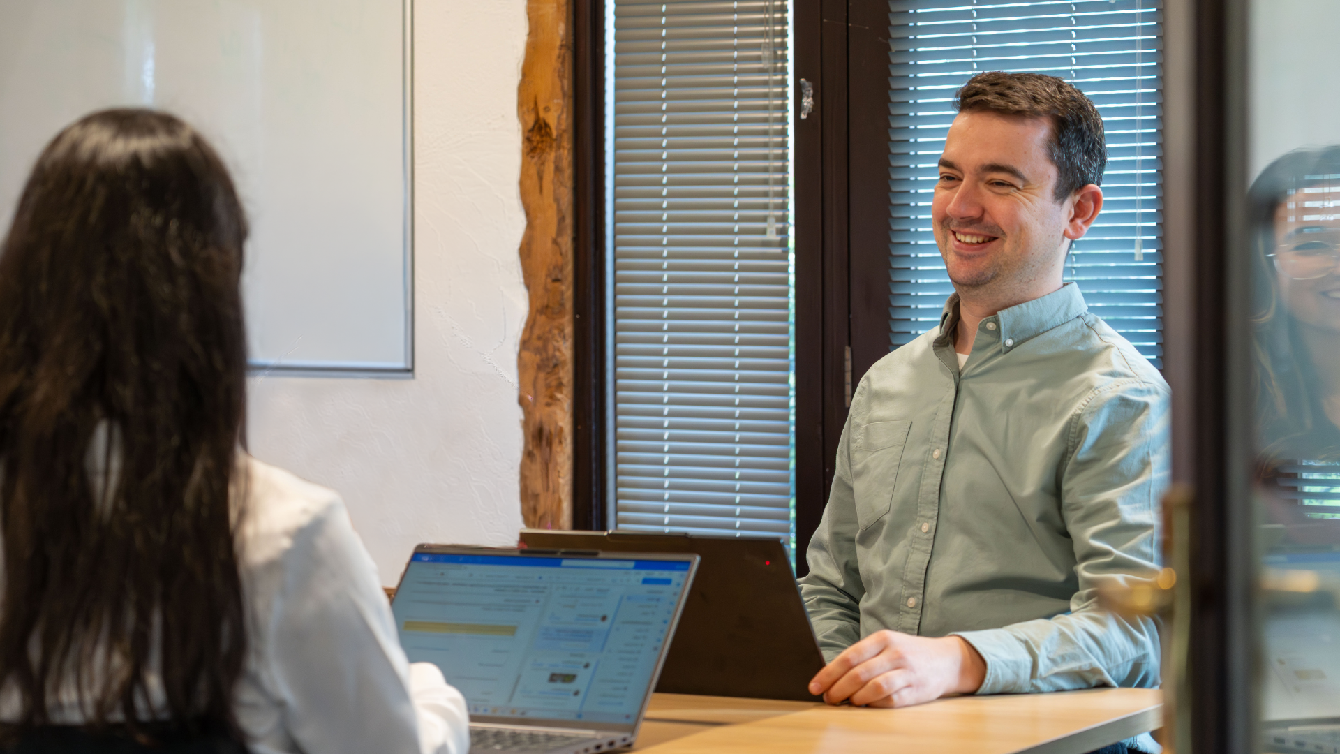 A man smiling during a conversation with a woman who is working on a laptop, seated at a table in a well-lit room with windows and blinds.