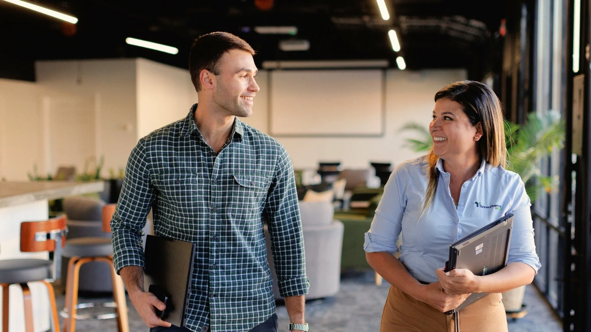 A man and a woman are walking and talking in an office or coworking space. The man wears a checkered shirt and holds a laptop, while the woman, smiling, wears a light blue button-up shirt with a logo and holds a laptop.