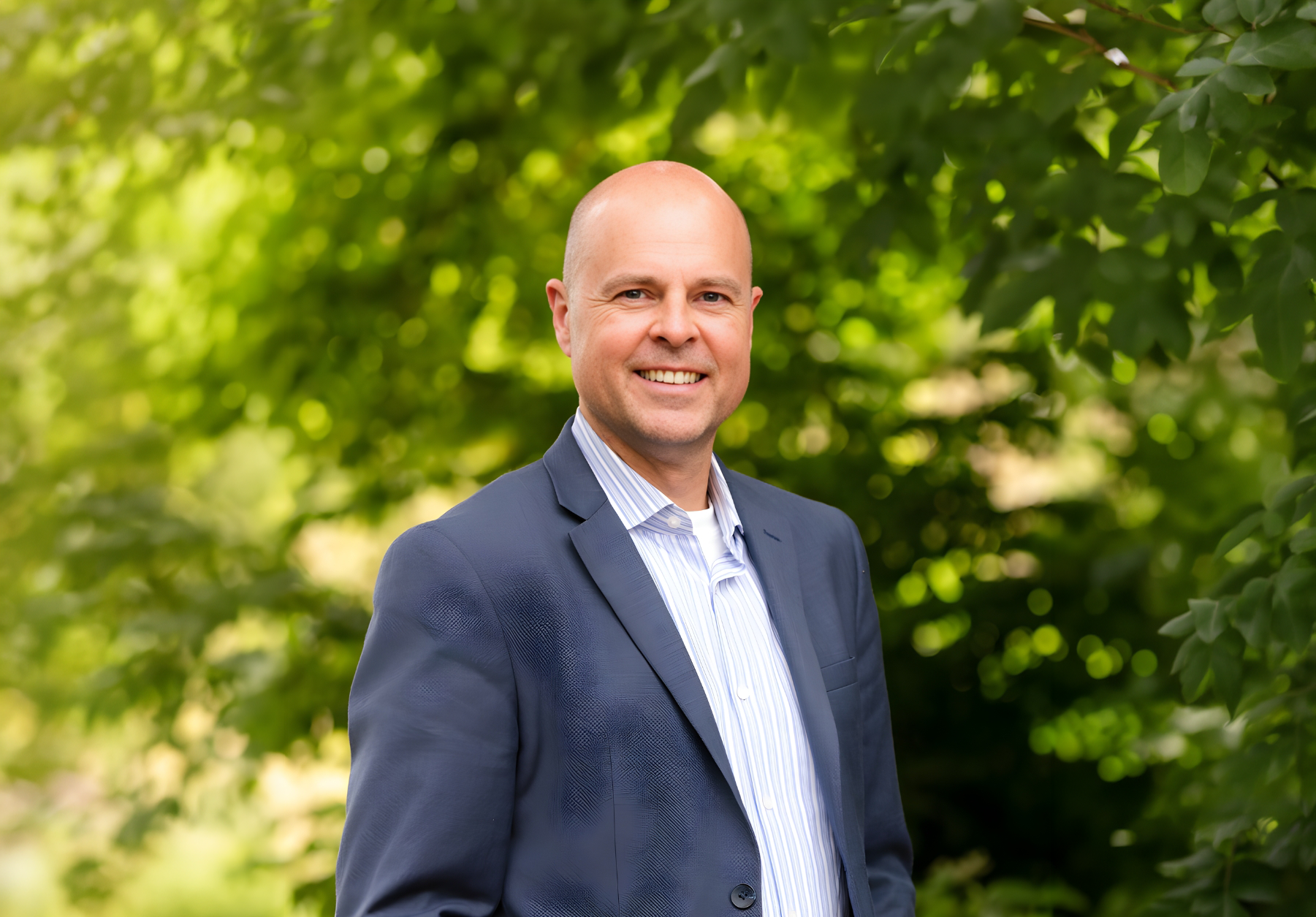A man in a suit smiling outdoors with green trees in the background.