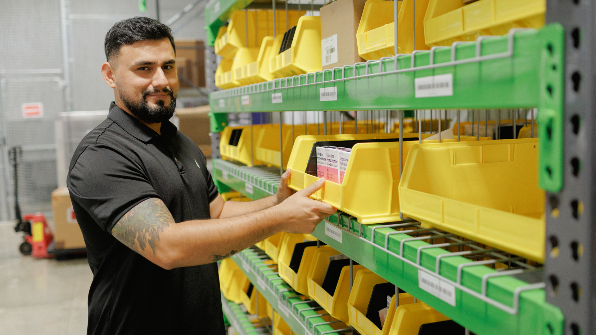 Man with a tattoo sleeve on his left arm looks at a yellow plastic storage bin filled with medical boxes on green shelving in a warehouse.