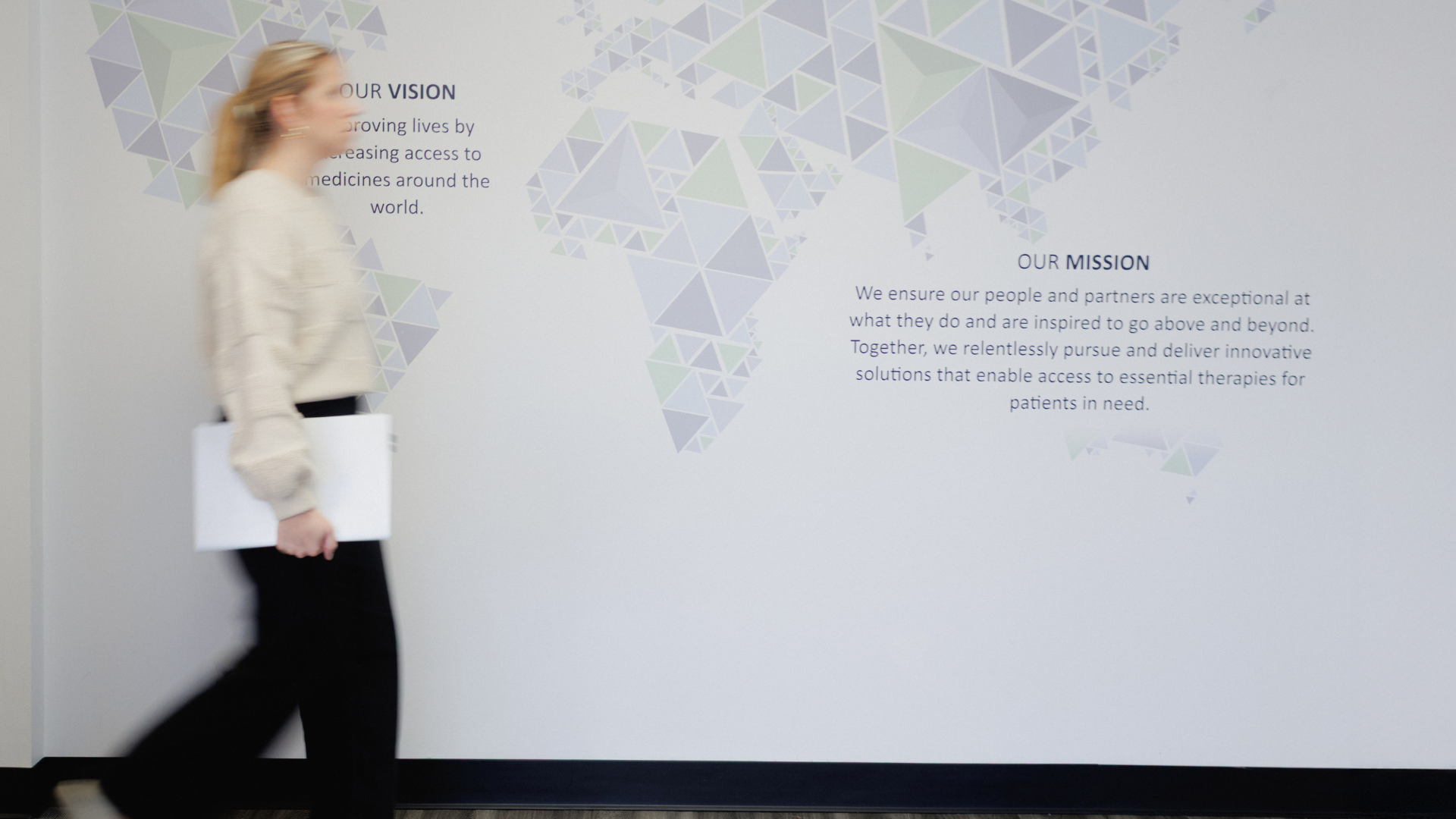 A woman walks past a wall with a world map and text about a company's vision and mission statements.
