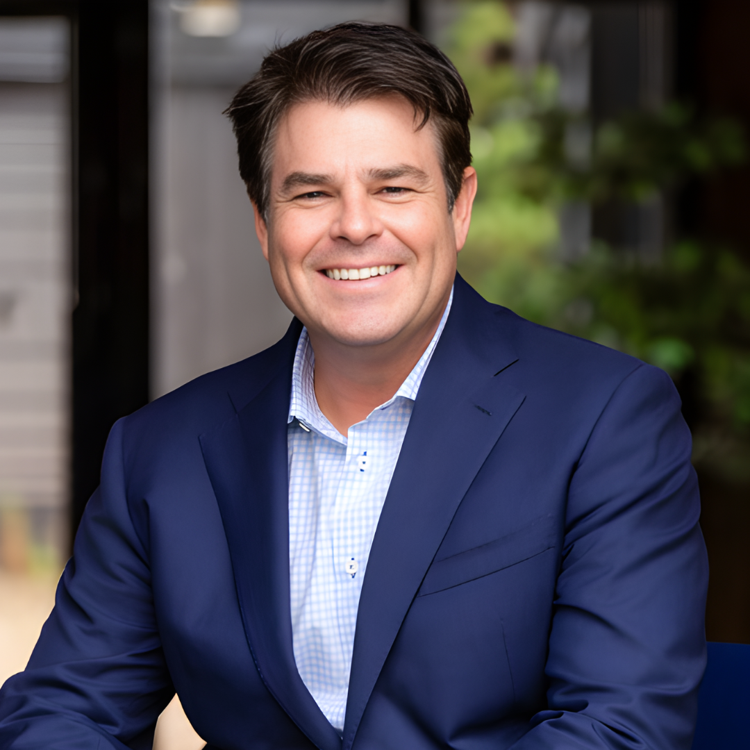 A smiling man in a blue suit and white shirt, sitting outdoors with greenery in the background.