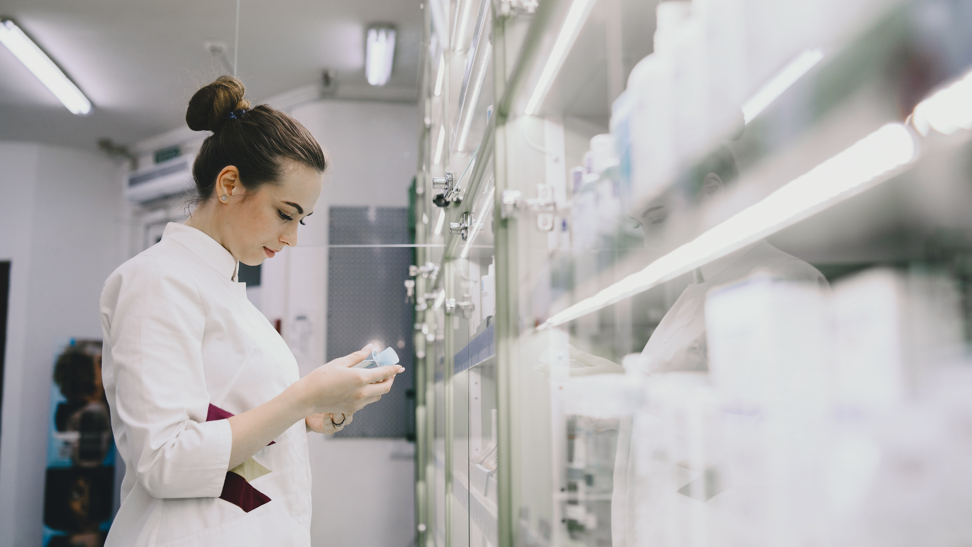 A female scientist in a white lab coat is examining a test tube in a laboratory with shelves of laboratory supplies in the background.