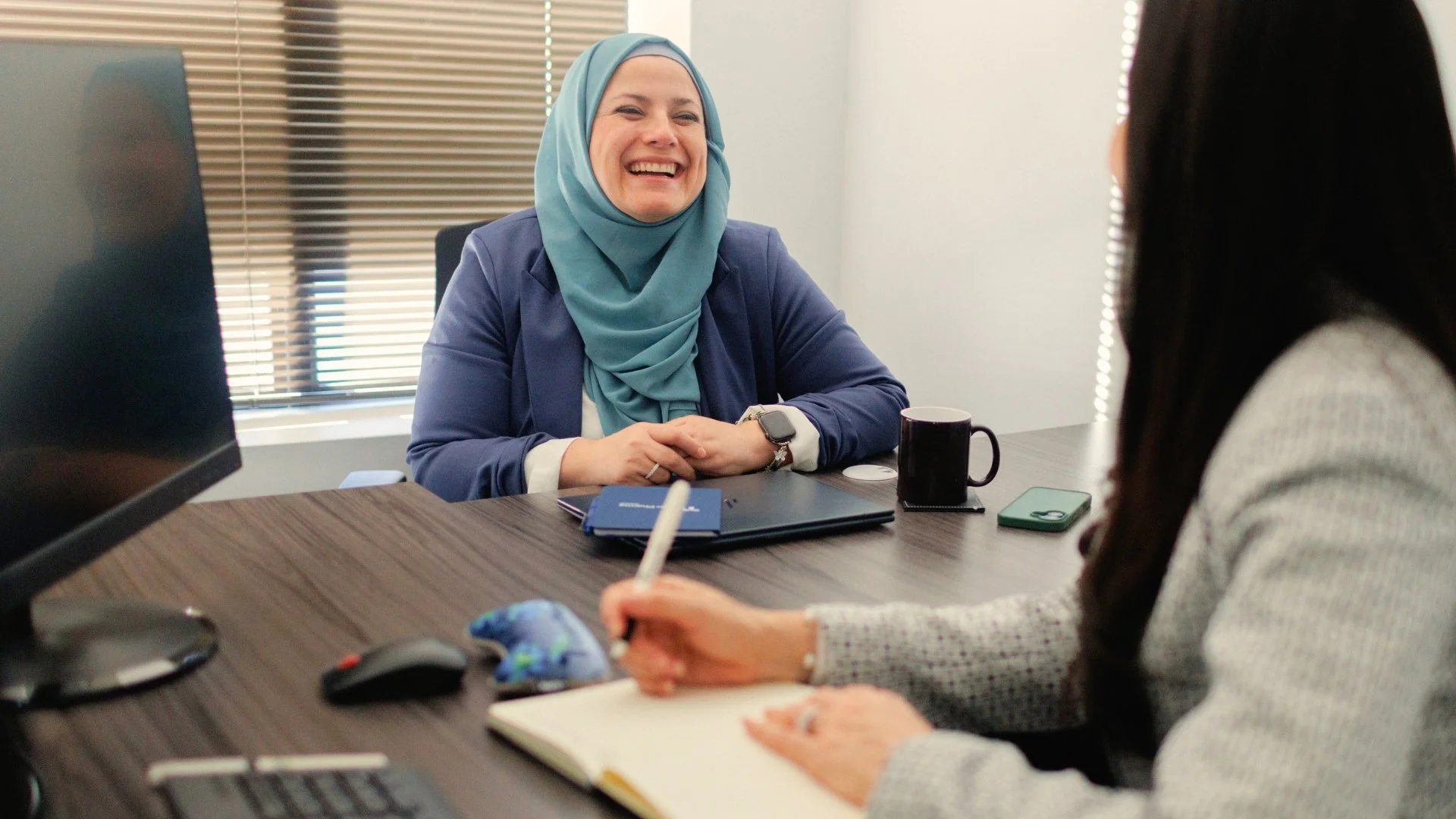 Two women having a professional meeting in an office. One woman, wearing a hijab and a blue blazer, is smiling and engaging in conversation. The other woman, with dark hair, is writing in a notebook.