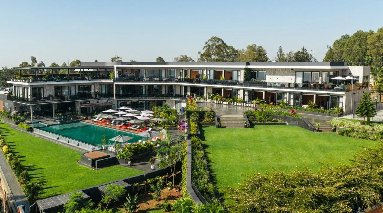 Modern multi-story hotel with large glass windows, outdoor pool with lounge chairs, umbrellas, and landscaped lawn, illuminated during dusk with a partly cloudy sky.