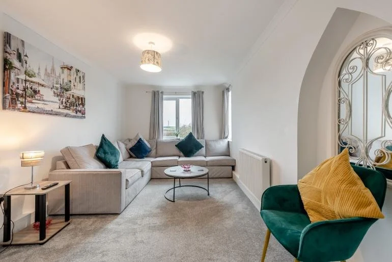 Living room with beige sofa, teal and navy cushions, a small round coffee table, a side table with a lamp, a wall art painting, and a green armchair with a yellow cushion, near arched window with ornate grill.