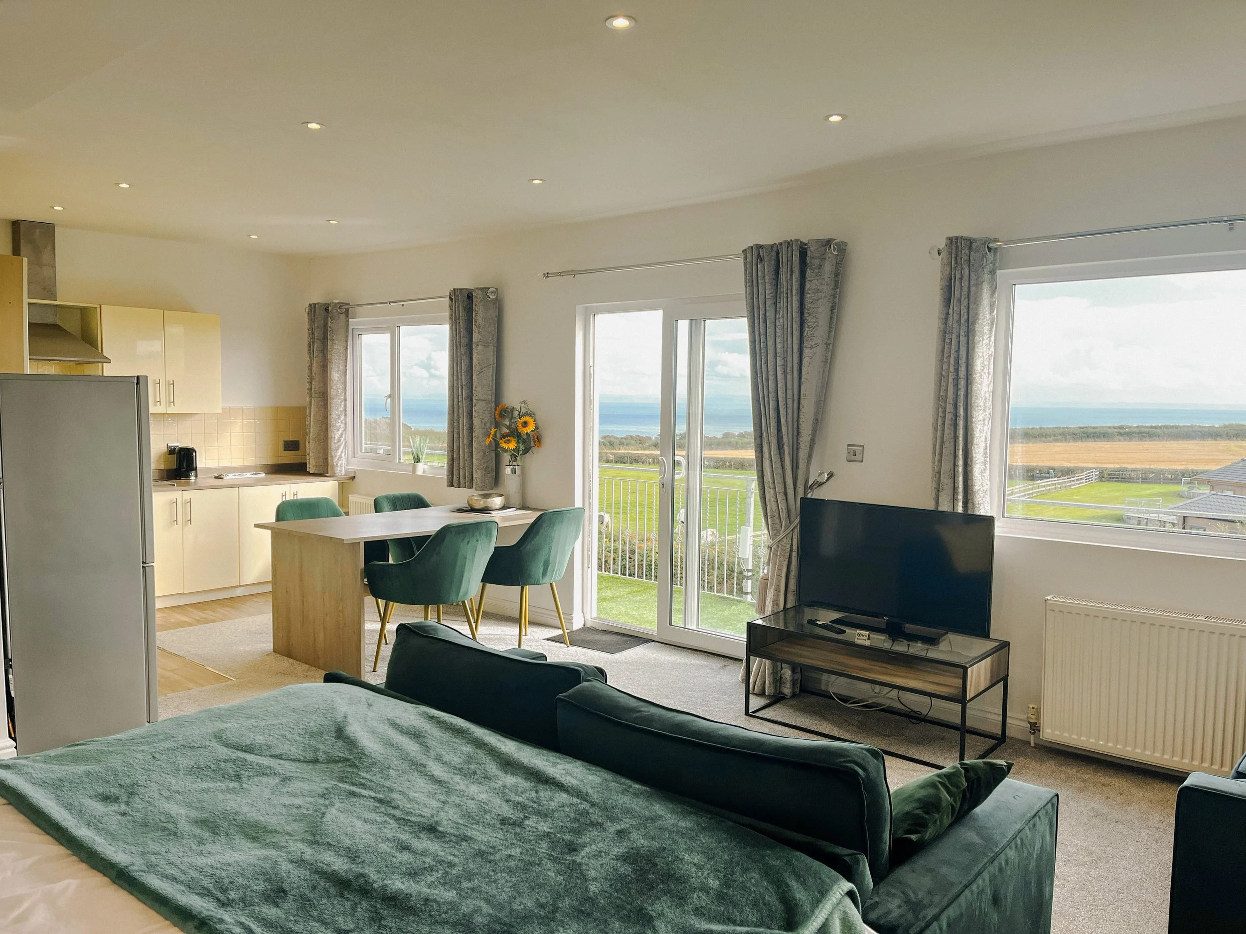 Living room with large windows and glass door overlooking a green landscape and ocean in the distance, with a bed, a TV on a stand, a dining table with four teal chairs, and kitchen appliances in the background.