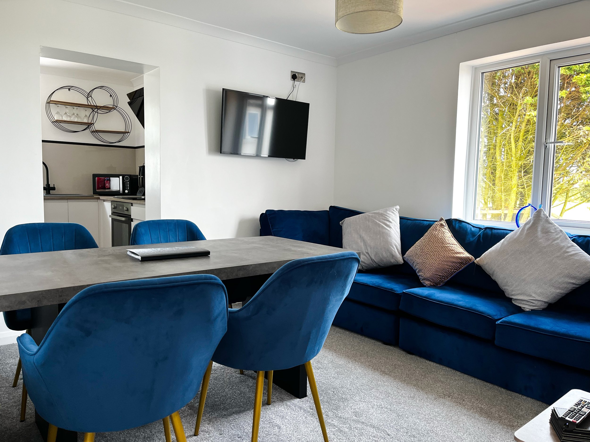 Dining area with a dark gray table set for breakfast, surrounded by blue chairs, with a blue sofa and large window in a bright, modern living space.