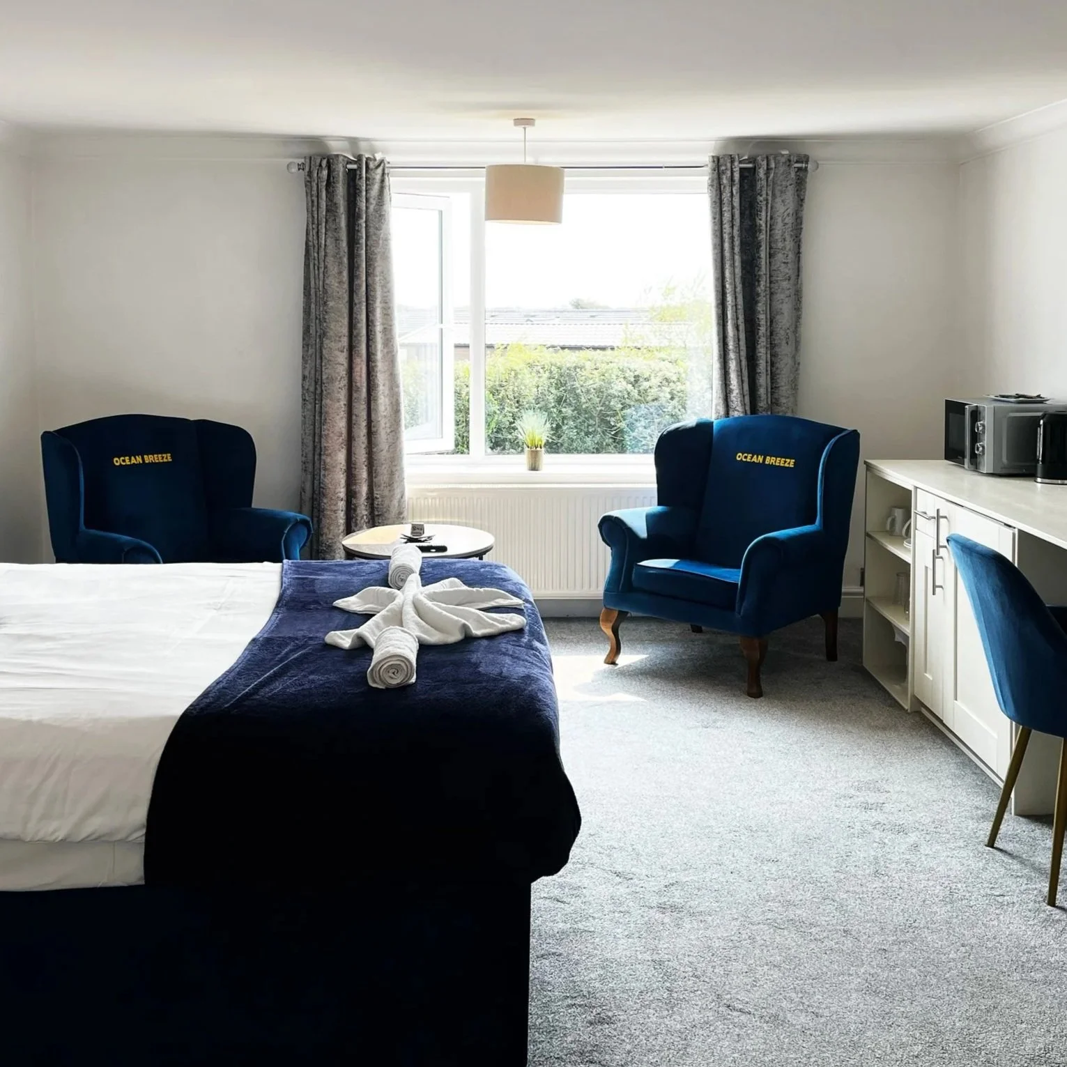 A hotel room with a bed, two blue armchairs with 'Ocean Breeze' embroidered on the back, a small round table, a window with curtains, and a white cabinet with a microwave and a cup.