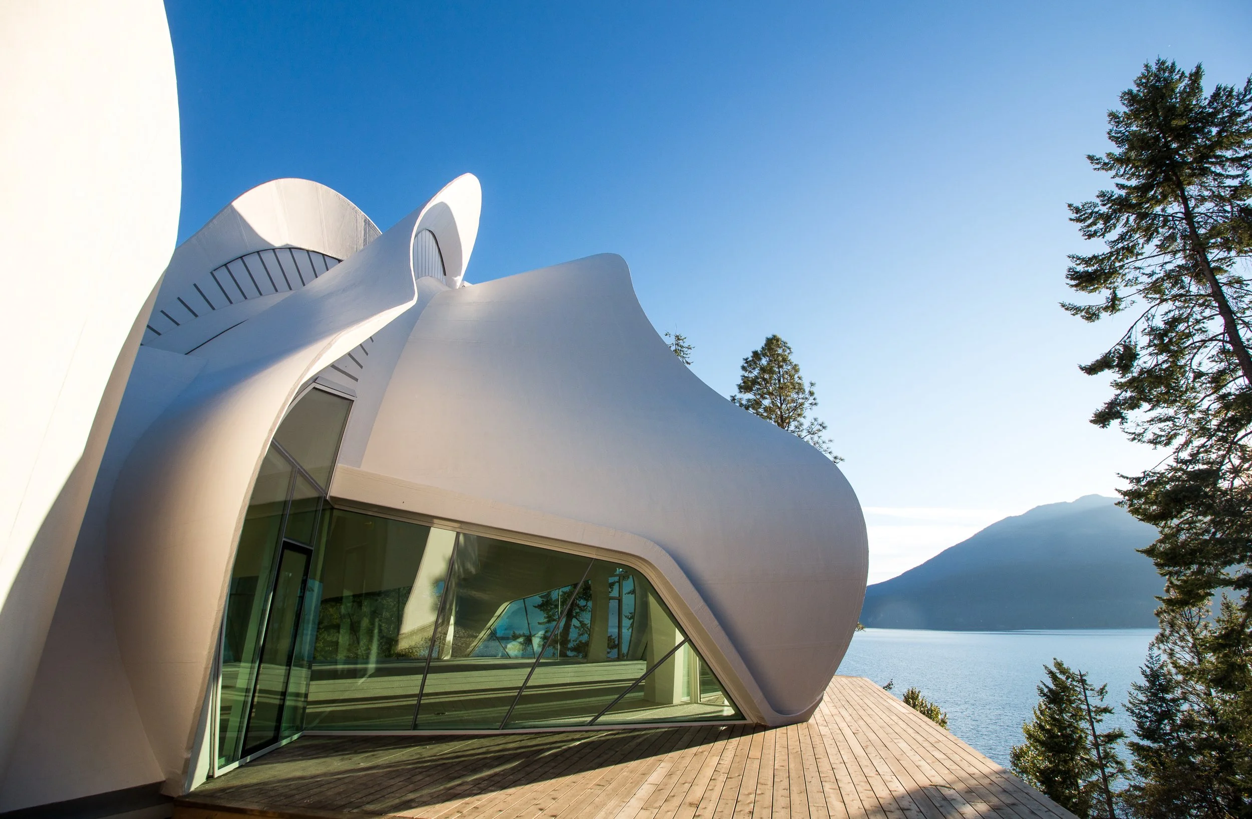Une architecture moderne en forme organique blanche, située au bord d'un lac avec des montagnes en arrière-plan et entourée d'arbres, sous un ciel clair.