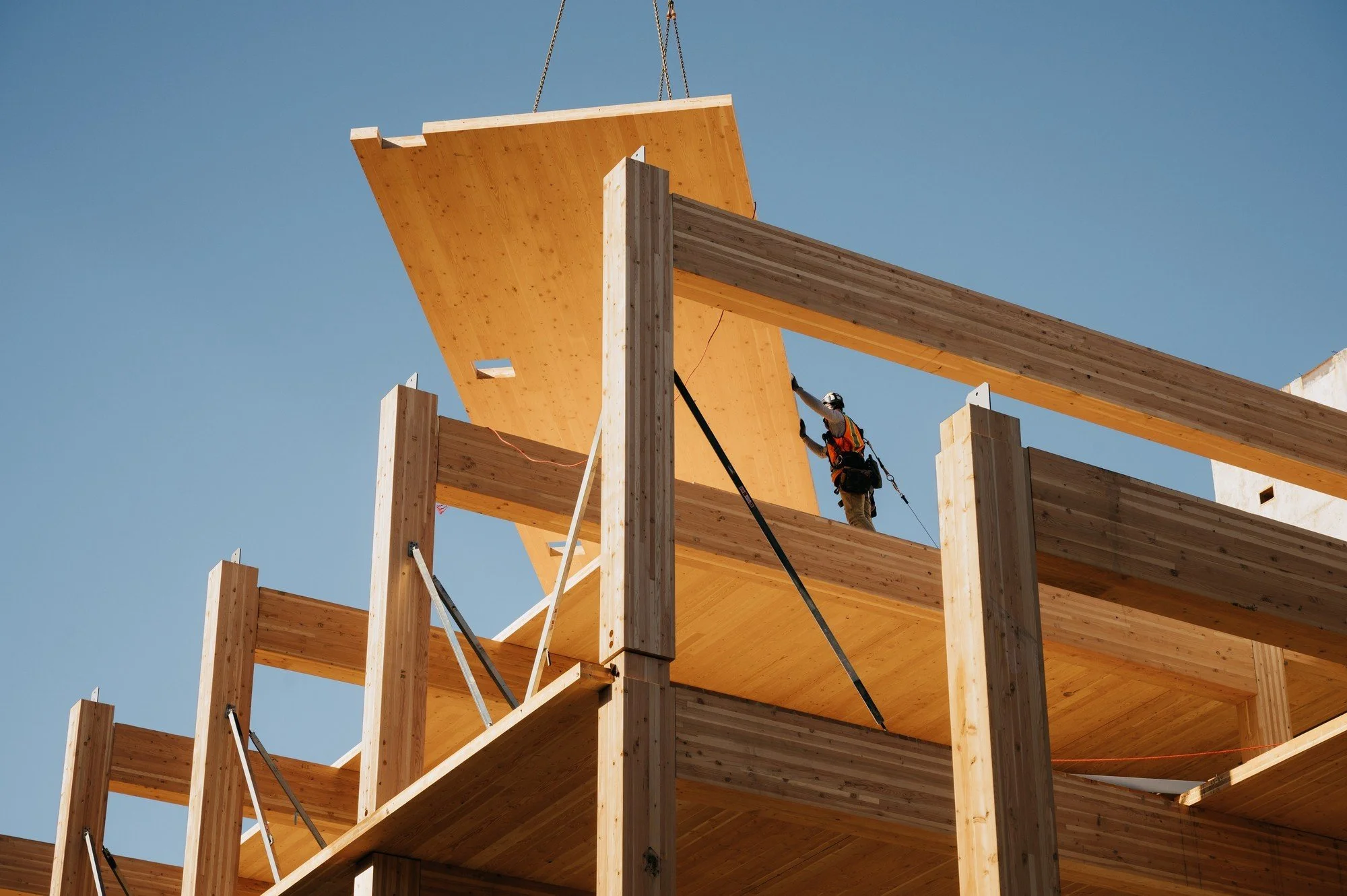 Un ouvrier travaille sur la construction d'un bâtiment en bois, en hauteur, avec plusieurs étages, sous un ciel clair.