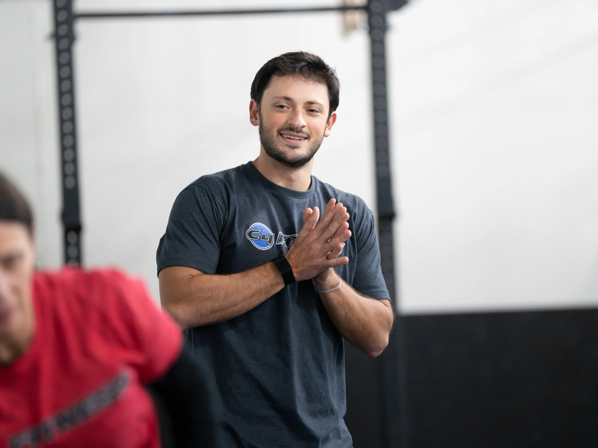 A man with dark hair and beard, wearing a fitness t-shirt, stands in a gym with his hands pressed together in a gesture of gratitude or greeting. In the foreground, another person wearing a red shirt is partially visible.
