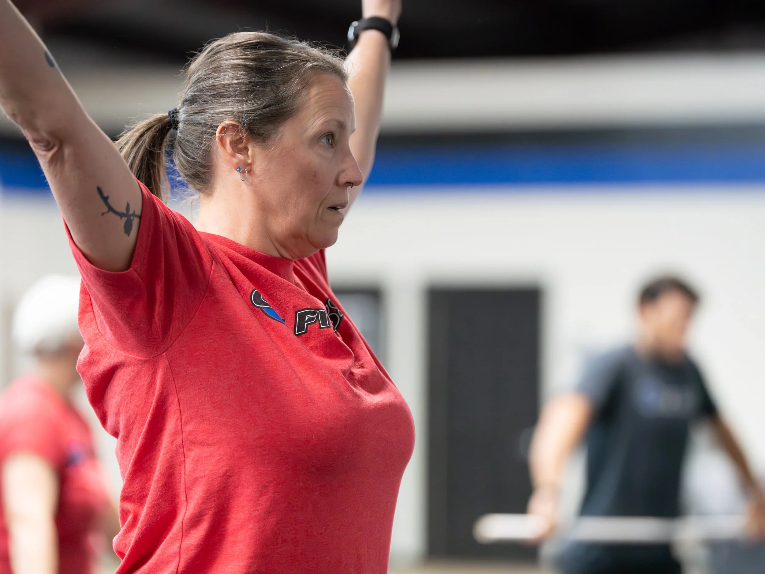 A woman in a red shirt stretching in a gym, with other people exercising blurred in the background.