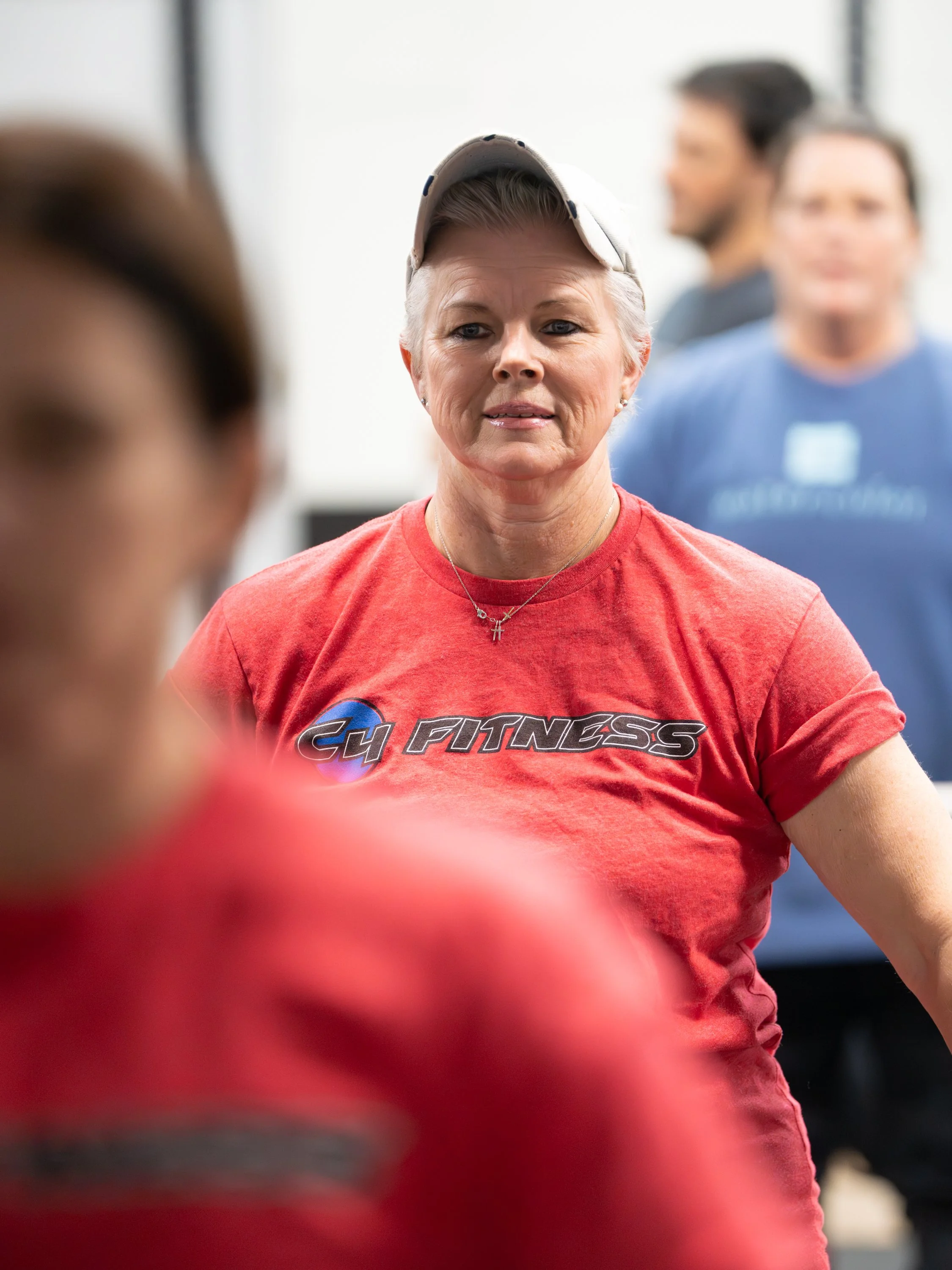 A woman with short gray hair wearing a red t-shirt with 'GH FITNESS' logo and a beige baseball cap. She is in a gym or fitness class setting with other people in the background.