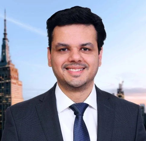 A man in a dark suit and tie smiling outdoors with city buildings in the background.