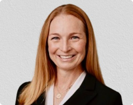 A smiling woman with long red hair wearing a black blazer and white blouse, standing against a white textured wall.